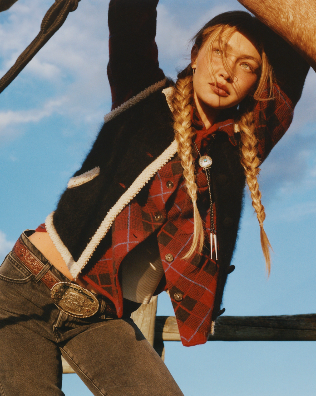 Woman with braided hair wearing a black jacket with cream trim over a red plaid shirt and jeans, posing outdoors beneath a bright blue sky.