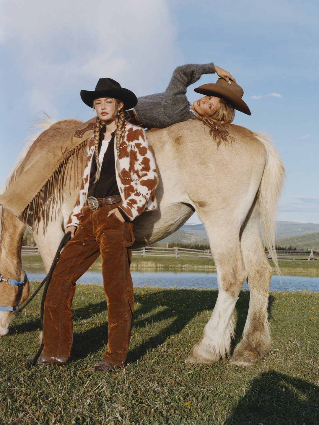 Two people in Western clothing pose beside a cream colored horse in a grassy field while one person leans across its back under a blue sky