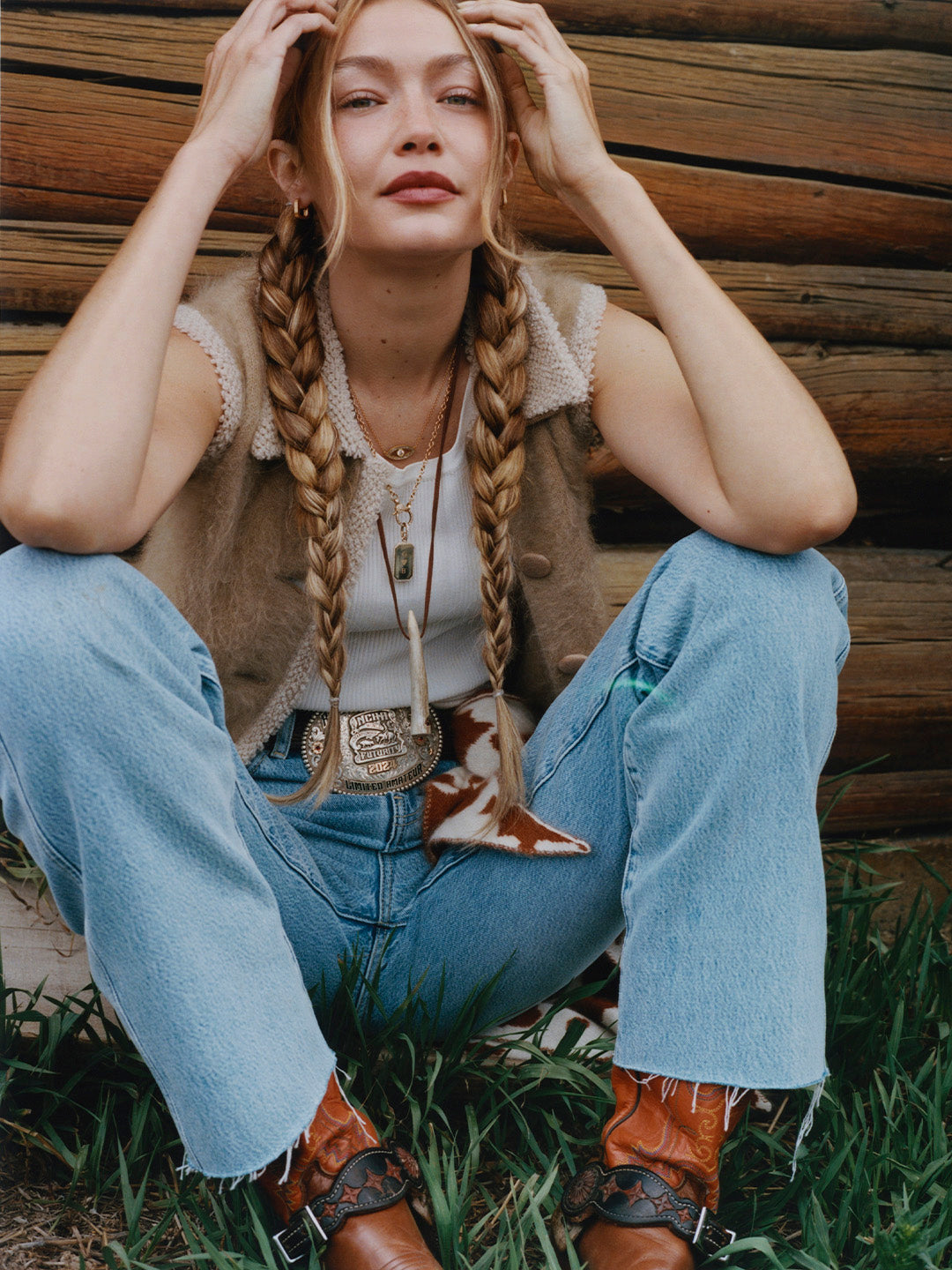 Woman with long braids sitting against a rustic wooden wall wearing a textured cream knit vest light blue jeans and embroidered brown boots