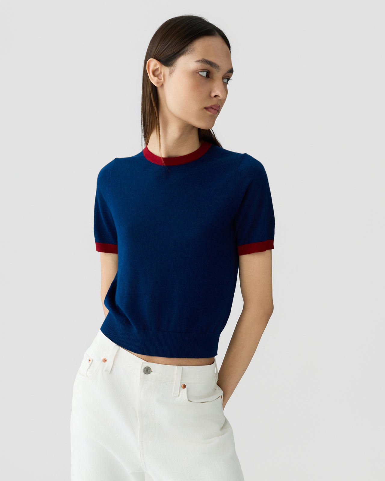 Young woman wearing a navy short-sleeve knit top with deep red trim at the collar and cuffs, paired with high-waisted white pants against a plain light background.