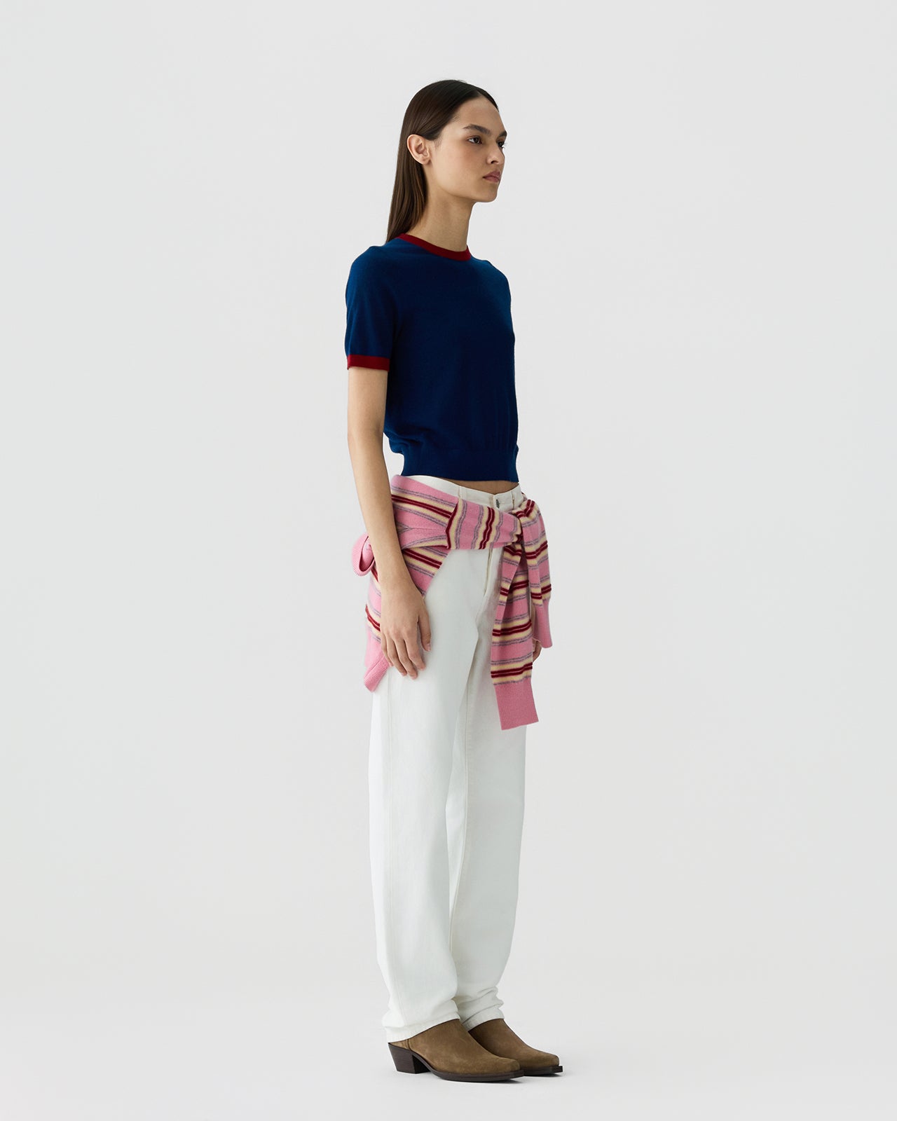 Woman standing in profile wearing a navy short-sleeve top with red trim at the collar and cuffs, white pants, and a pink striped sweater tied around her waist paired with brown boots.