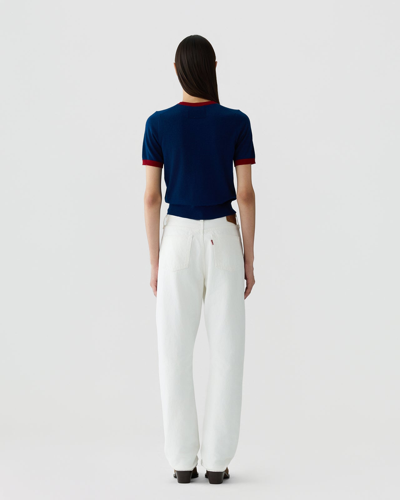 Woman standing facing away wearing a navy short-sleeve fitted top with contrasting red trim on collar and cuffs, paired with high-waisted white wide-leg pants against a plain background.