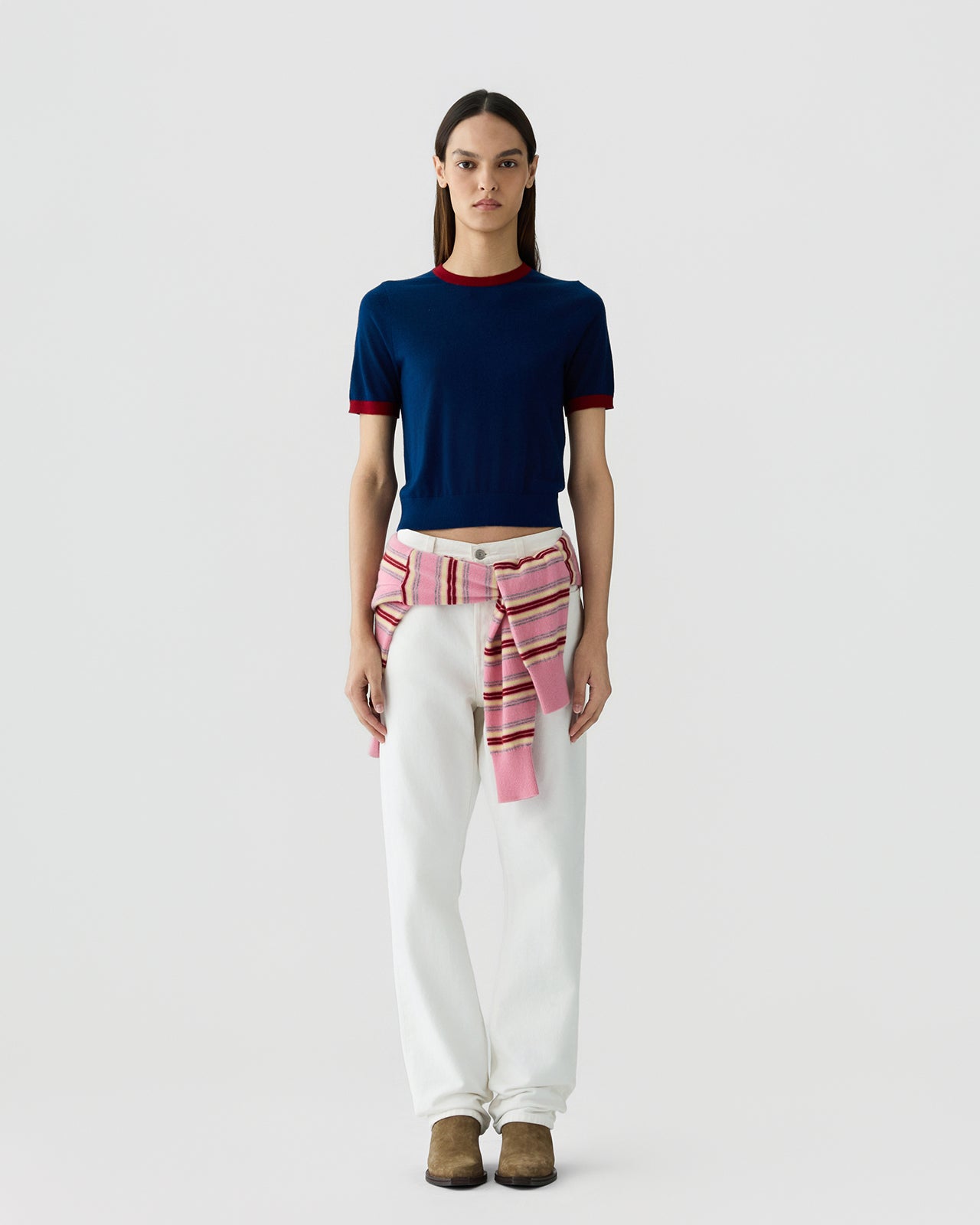 Woman standing facing forward wearing a navy short-sleeve fitted top with deep red trim at the collar and sleeves, paired with white pants and a pink striped sweater tied around her waist, brown shoes.