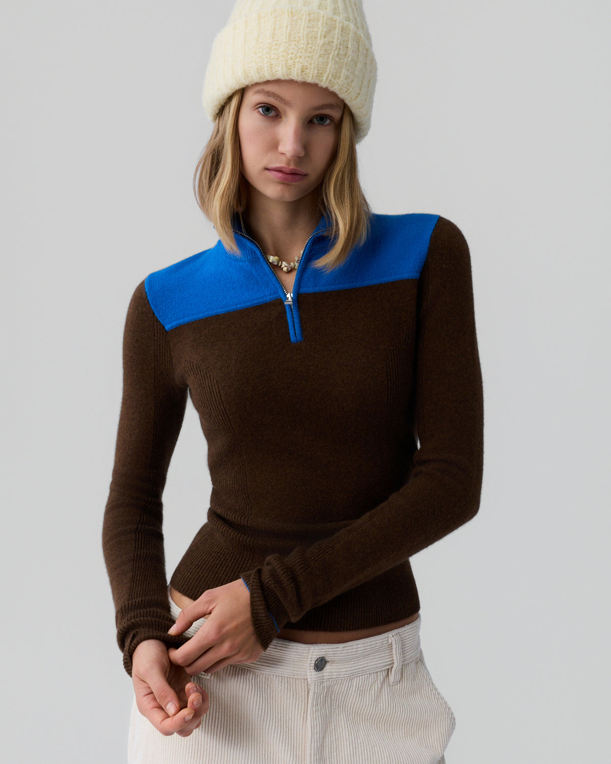 Young woman facing forward wearing a fitted chestnut brown half-zip sweater with a bright blue shoulder yoke, light-colored ribbed pants, and a cream knit beanie against a plain background.