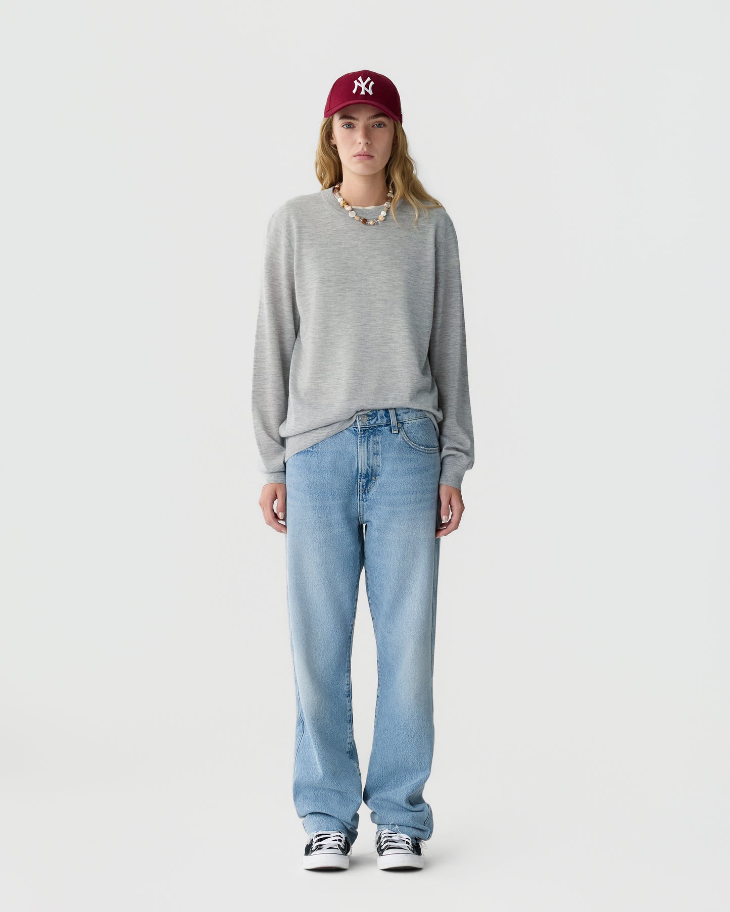 Person wearing a gray long sleeve sweater light blue jeans and a burgundy baseball cap standing against a plain light gray background.