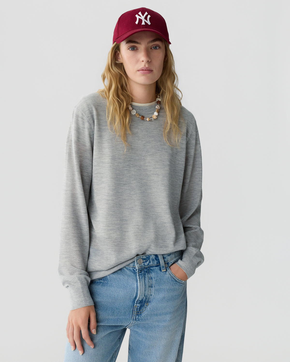 Woman with long wavy hair wearing a gray long sleeve sweater blue jeans and a burgundy baseball cap standing against a light gray background