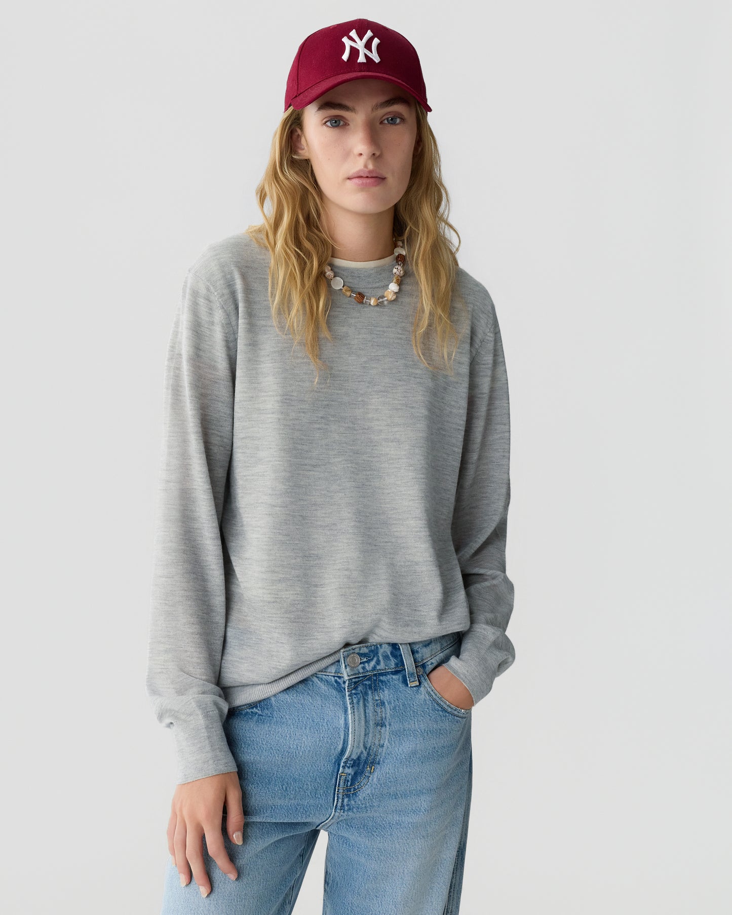 Woman with long wavy hair wearing a gray long sleeve sweater blue jeans and a burgundy baseball cap standing against a light gray background