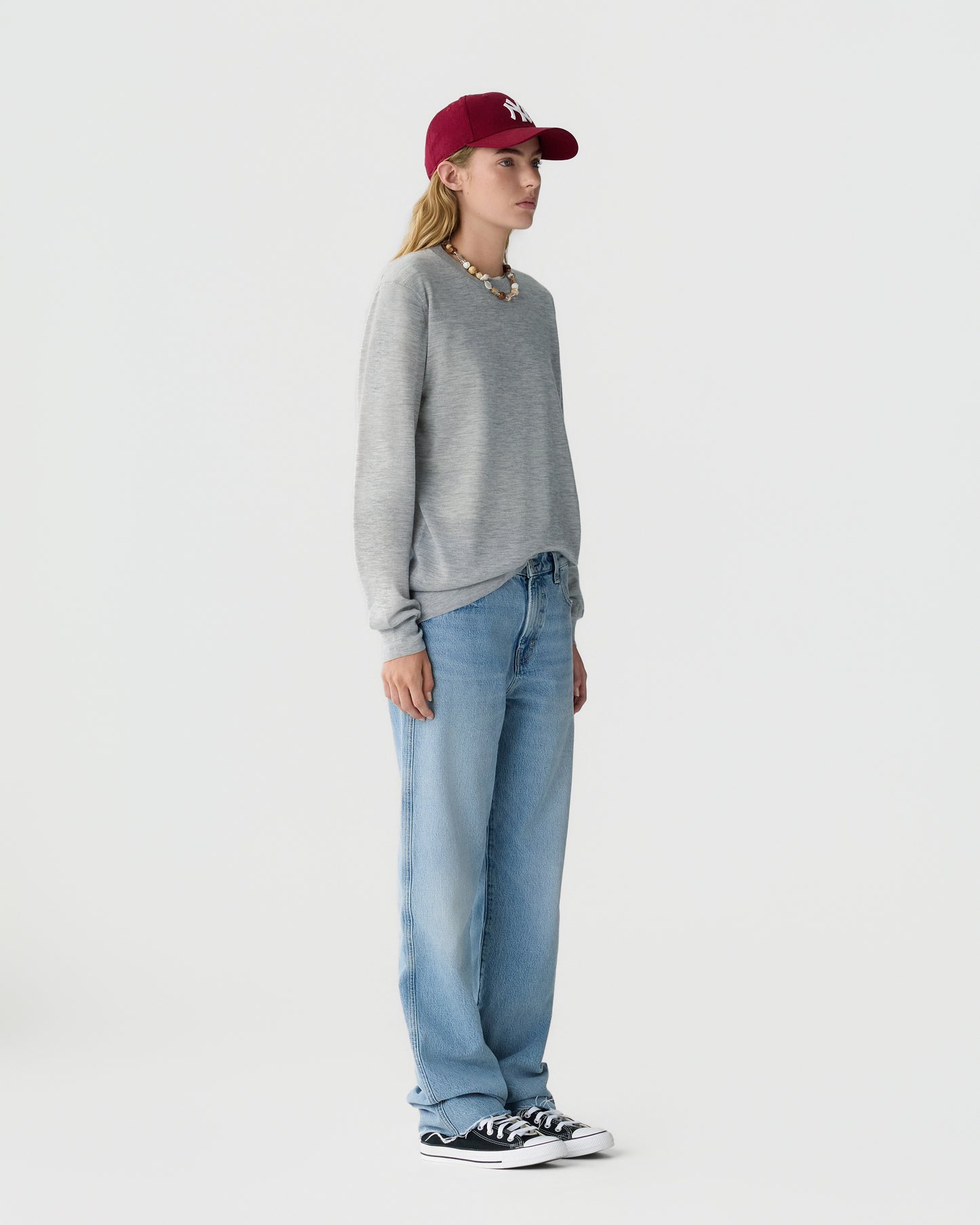 Person wearing a gray crewneck sweater, loose blue jeans, black and white sneakers, and a burgundy baseball cap against a light gray background