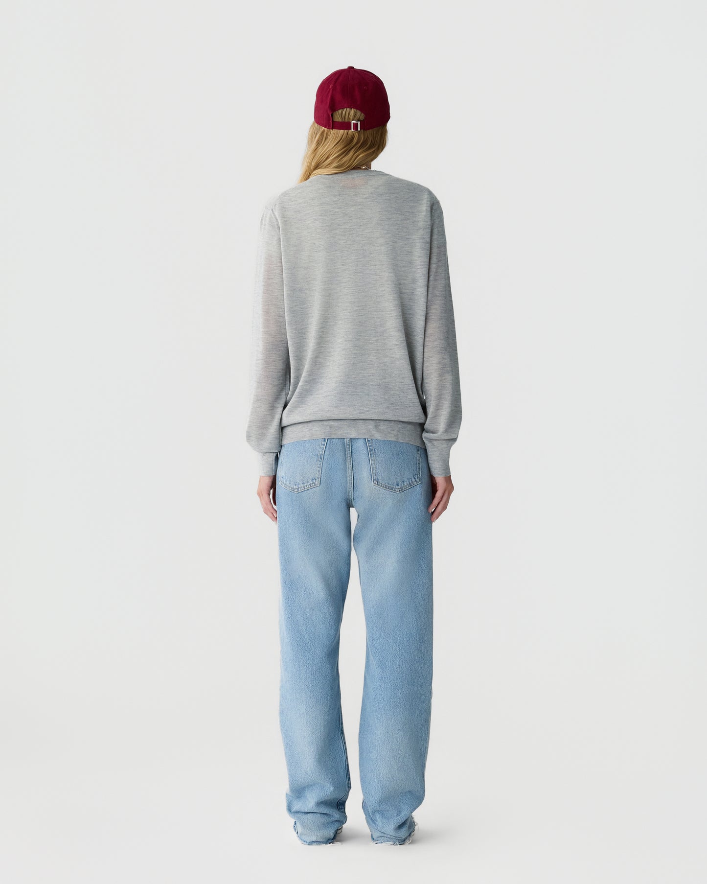 Back view of a person wearing a gray sweater, light blue jeans, and a red beanie against a plain white background