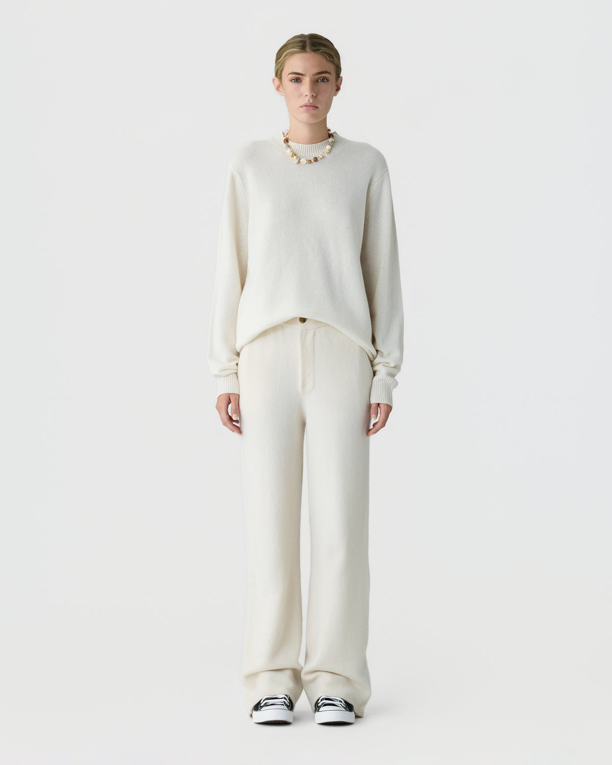 Model wearing an ivory cashmere sweater with relaxed cream trousers and black and white sneakers against a light gray studio background