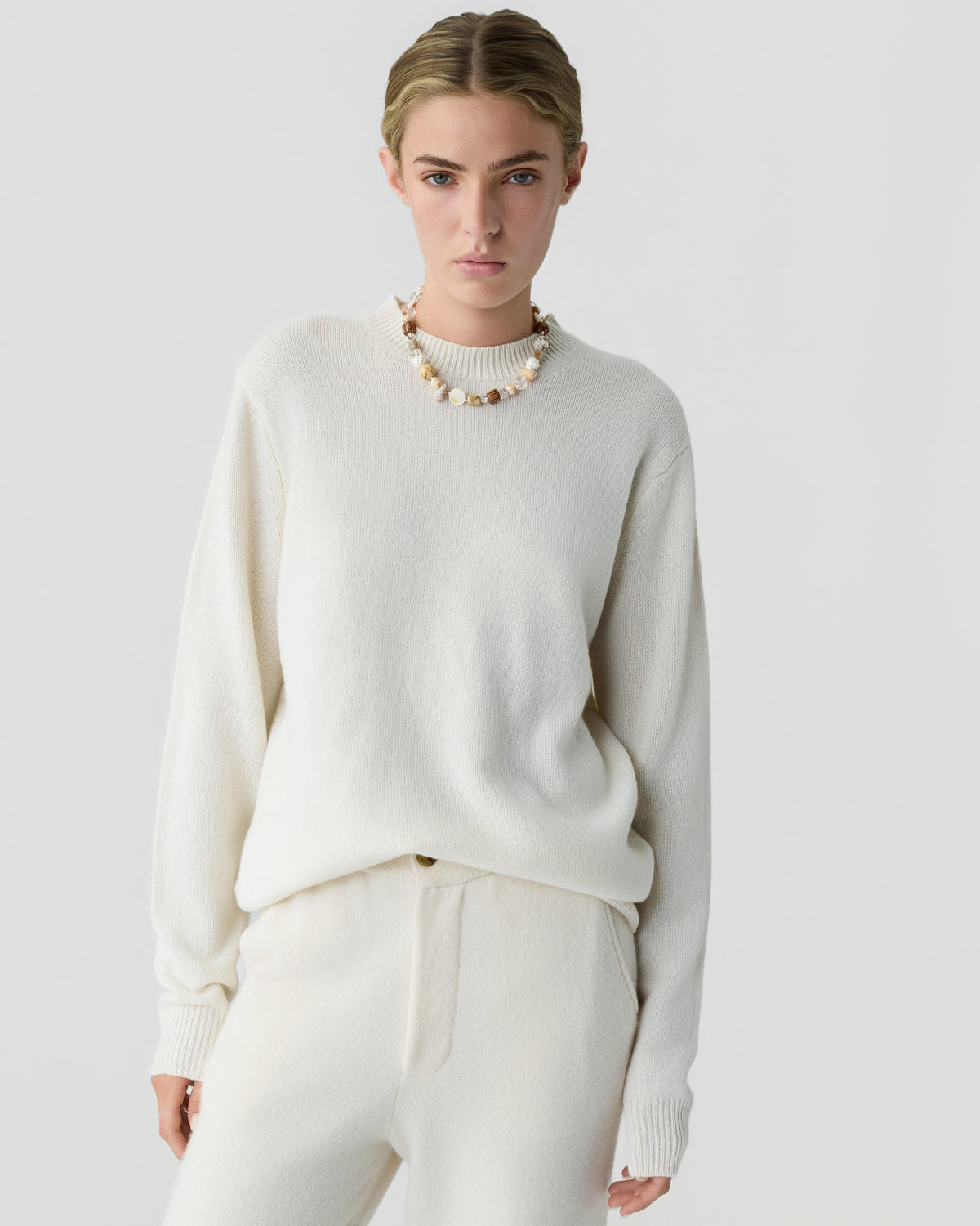 Model wearing an ivory cashmere sweater with ribbed cuffs and matching relaxed trousers accessorized with a beaded necklace against a light gray background