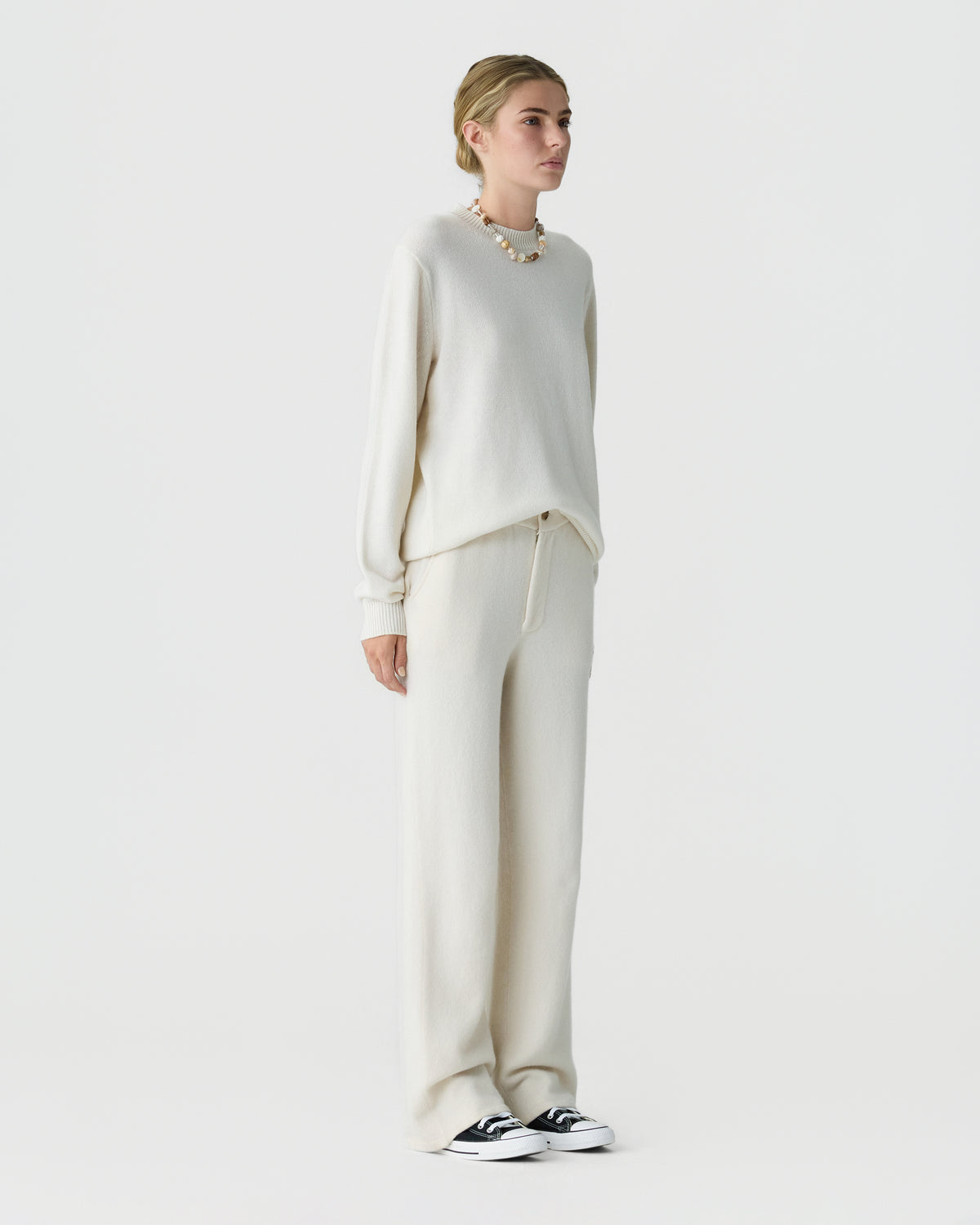 Full length view of a woman wearing a cream knit sweater with matching wide leg trousers a beaded necklace and black and white sneakers against a light background