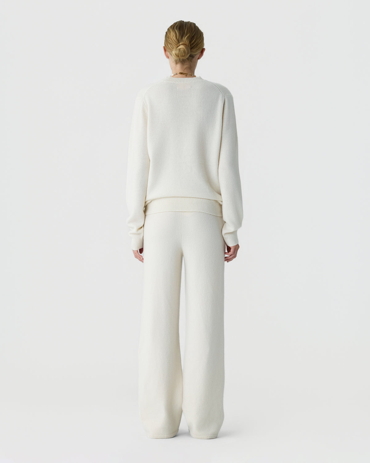 Back view of a person wearing a cream knit sweater and matching wide leg trousers, standing against a plain light background.