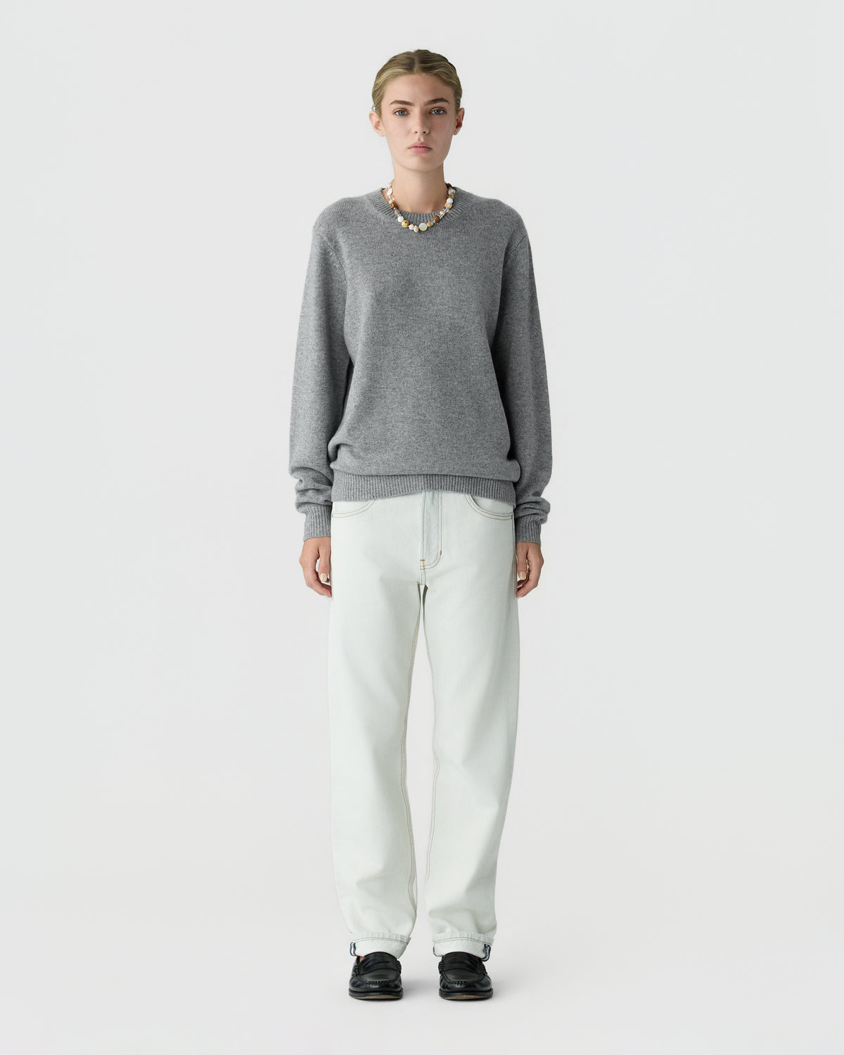 Full length view of a model wearing a gray crew neck sweater with rolled cuffs white straight leg trousers and black shoes against a light gray background
