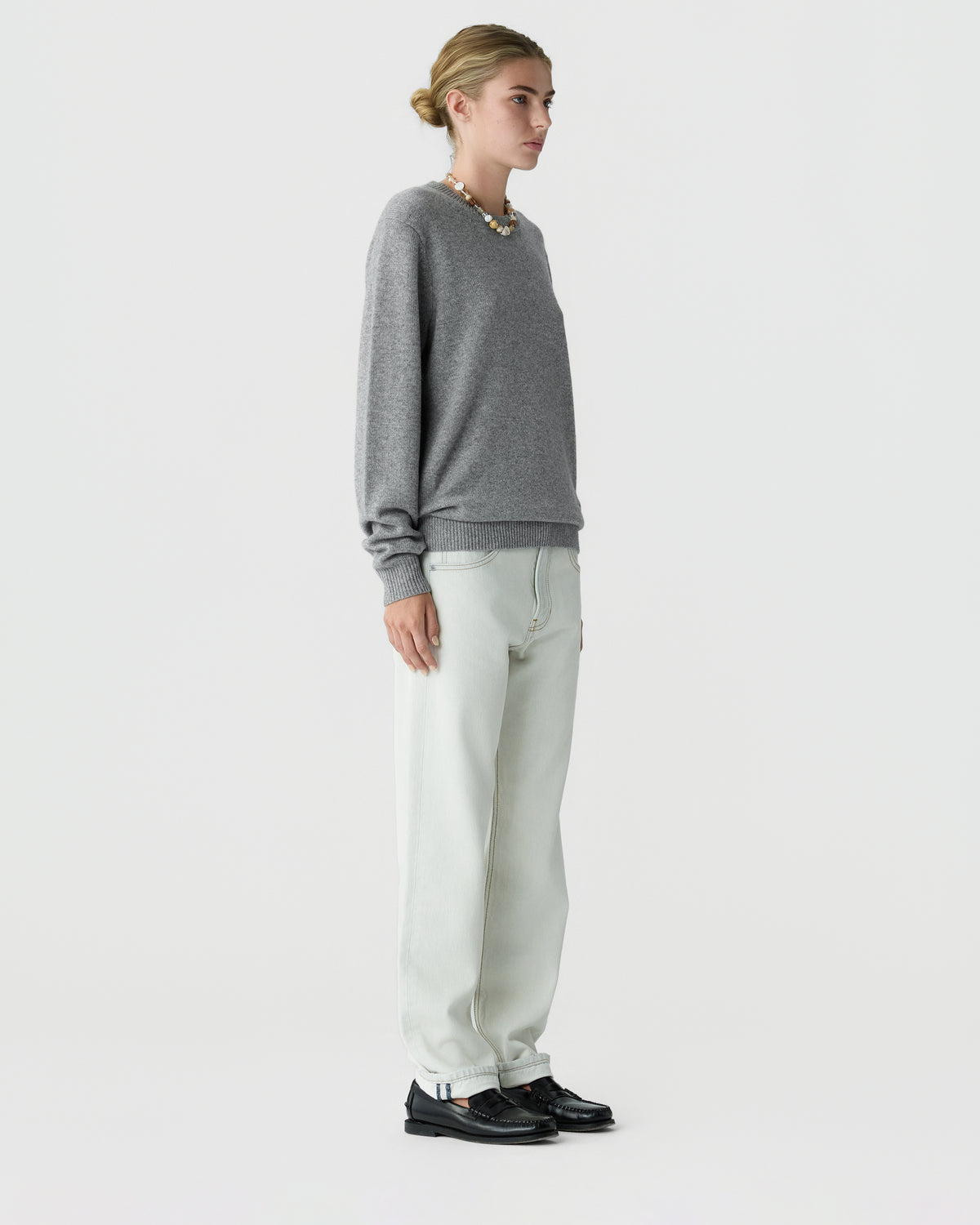 Woman in a gray crew neck sweater, light trousers, and black shoes standing in profile against a plain light background.