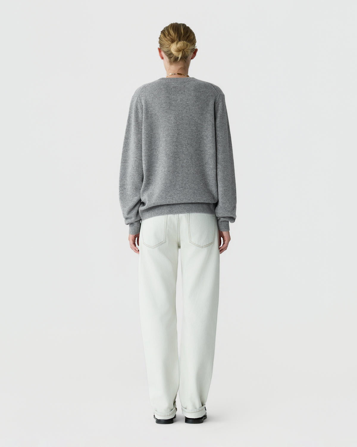 Person seen from behind wearing a gray crew neck sweater, white trousers, and black shoes against a plain light background.