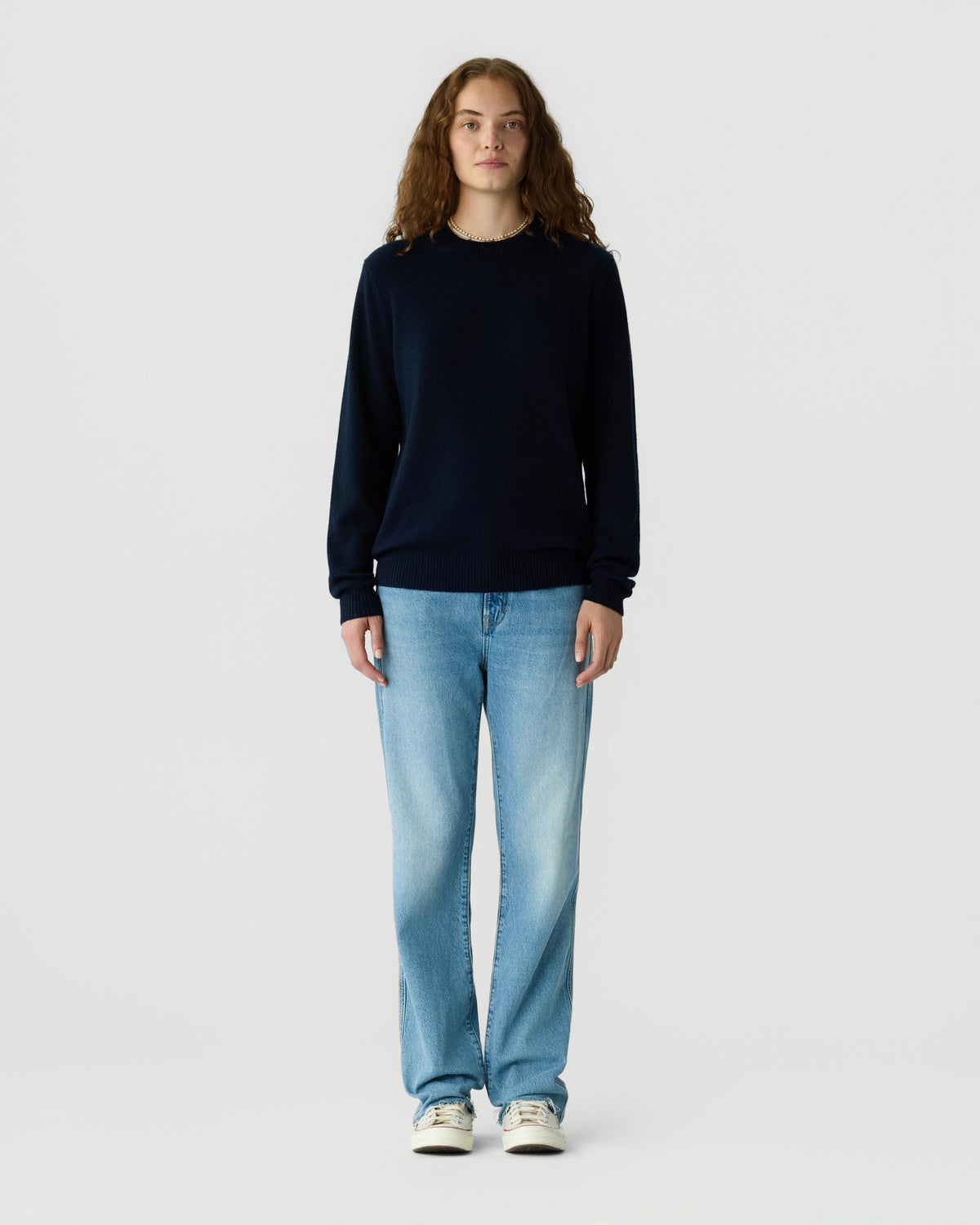Person with long brown hair wearing a dark navy crewneck sweater, light blue jeans, and white sneakers against a plain light gray background