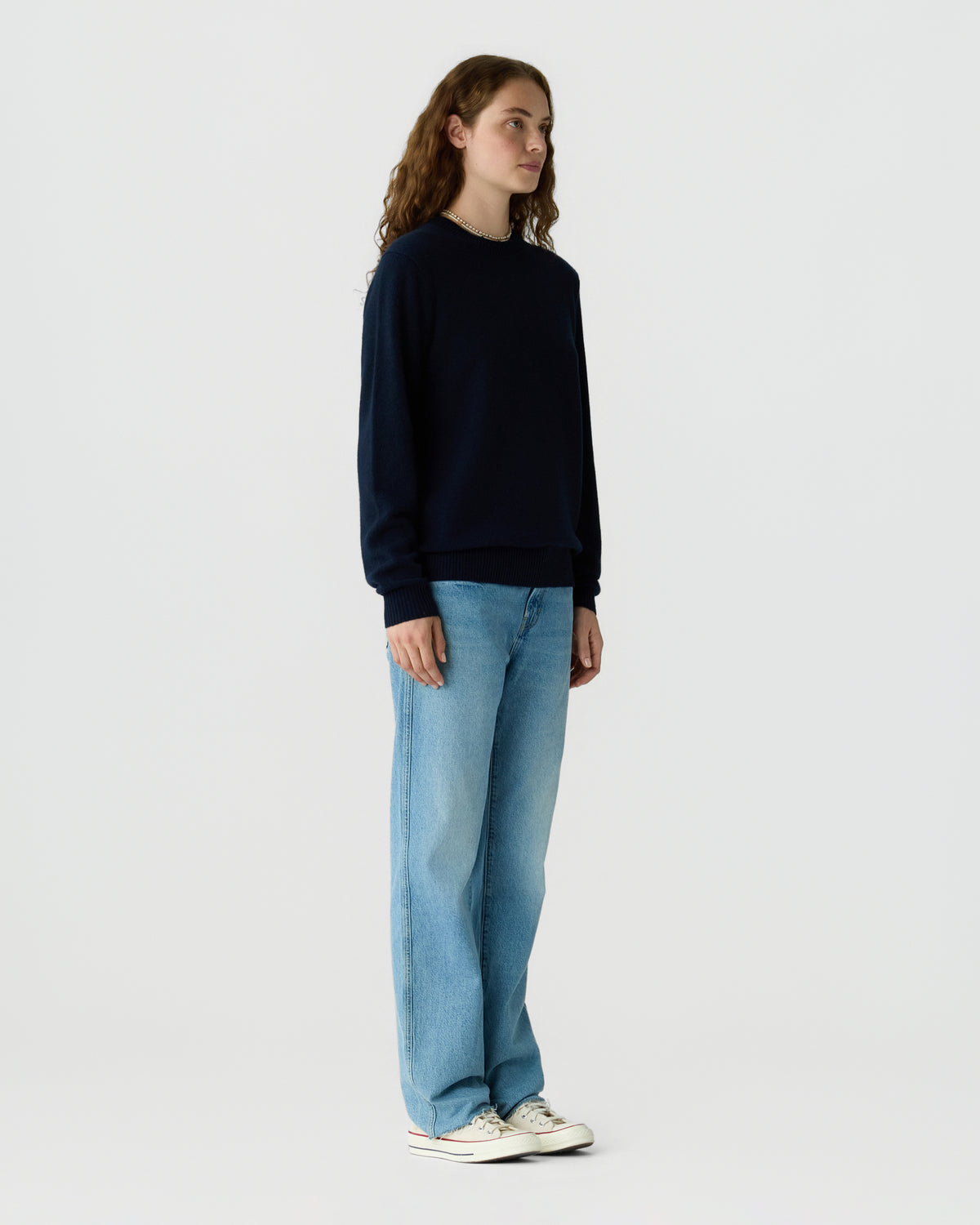 Woman standing in a navy crew neck sweater, light blue jeans, and white sneakers against a plain white studio background