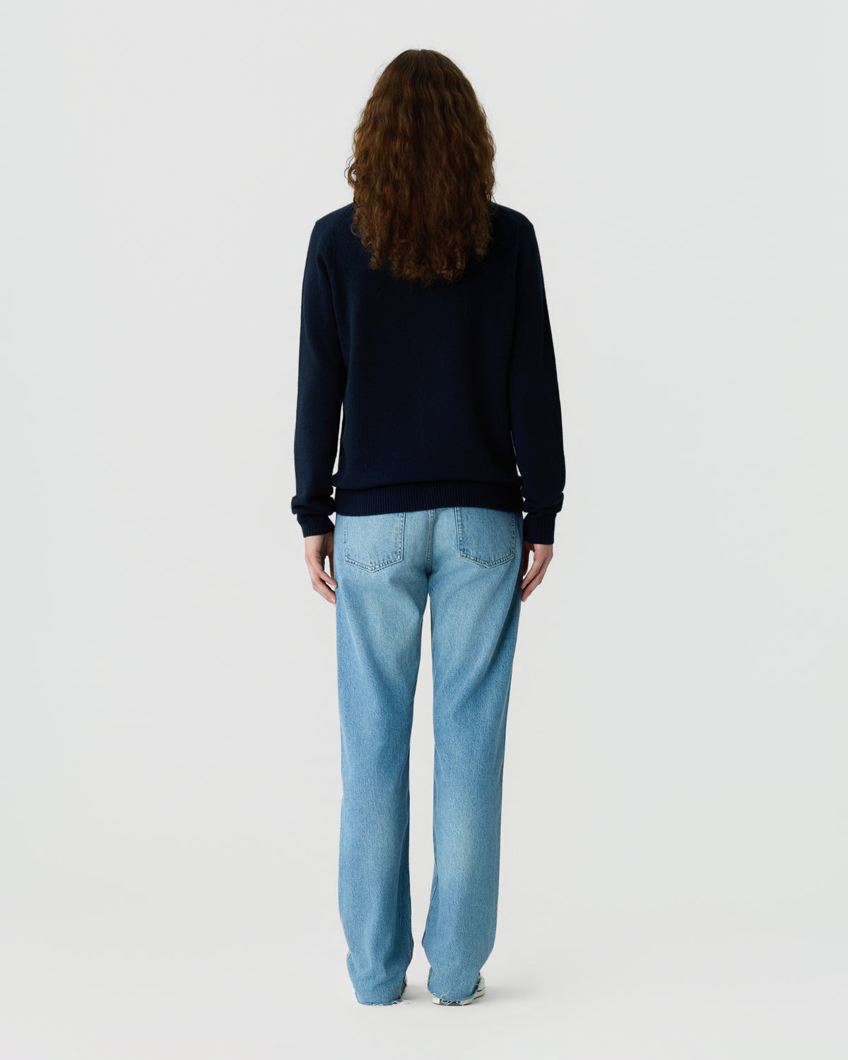 Back view of a person with long curly brown hair wearing a navy sweater and light blue jeans against a plain light gray background