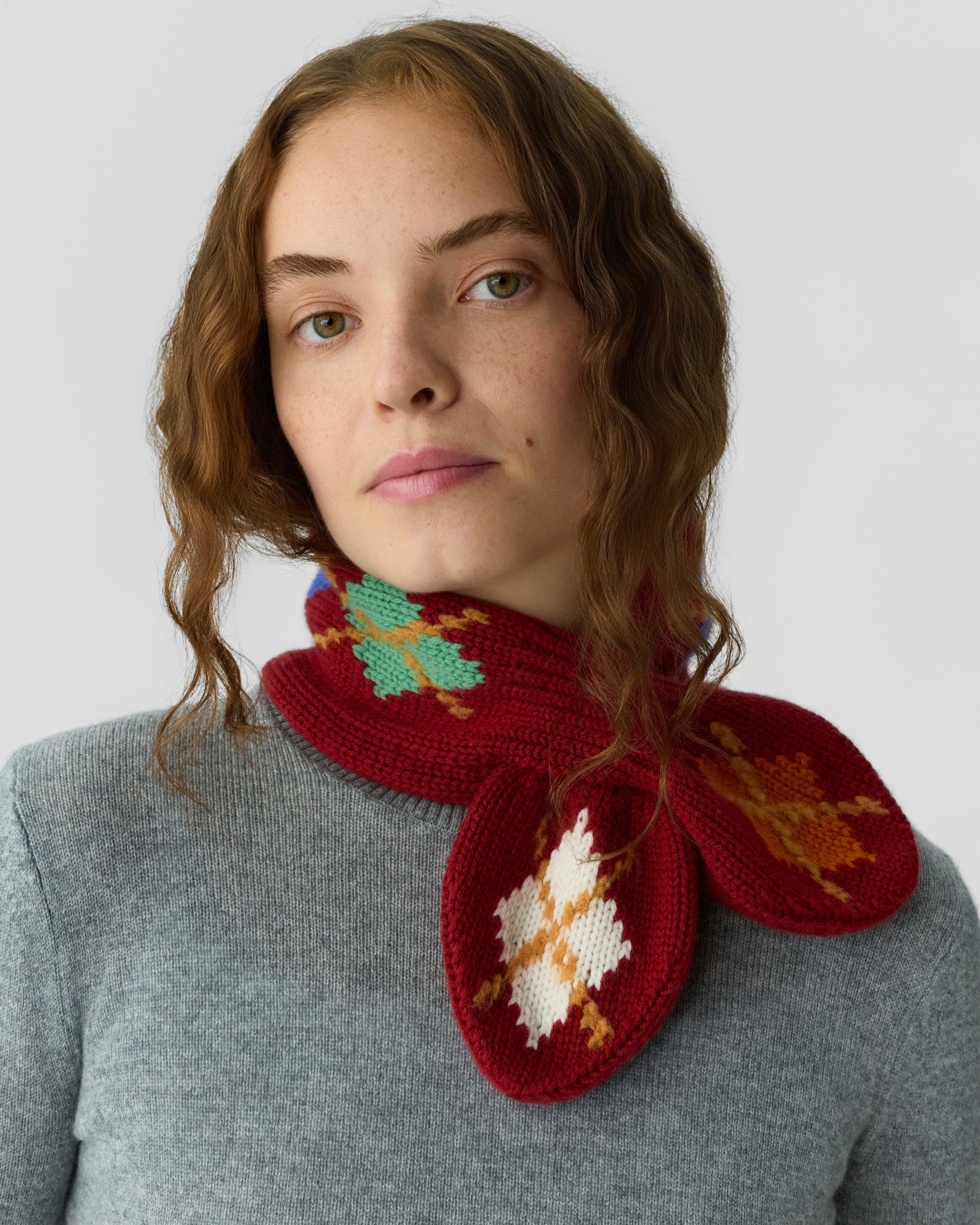 Young woman with wavy brown hair wearing a gray sweater and a red knitted scarf featuring colorful leaf-like patterns wrapped around her neck.