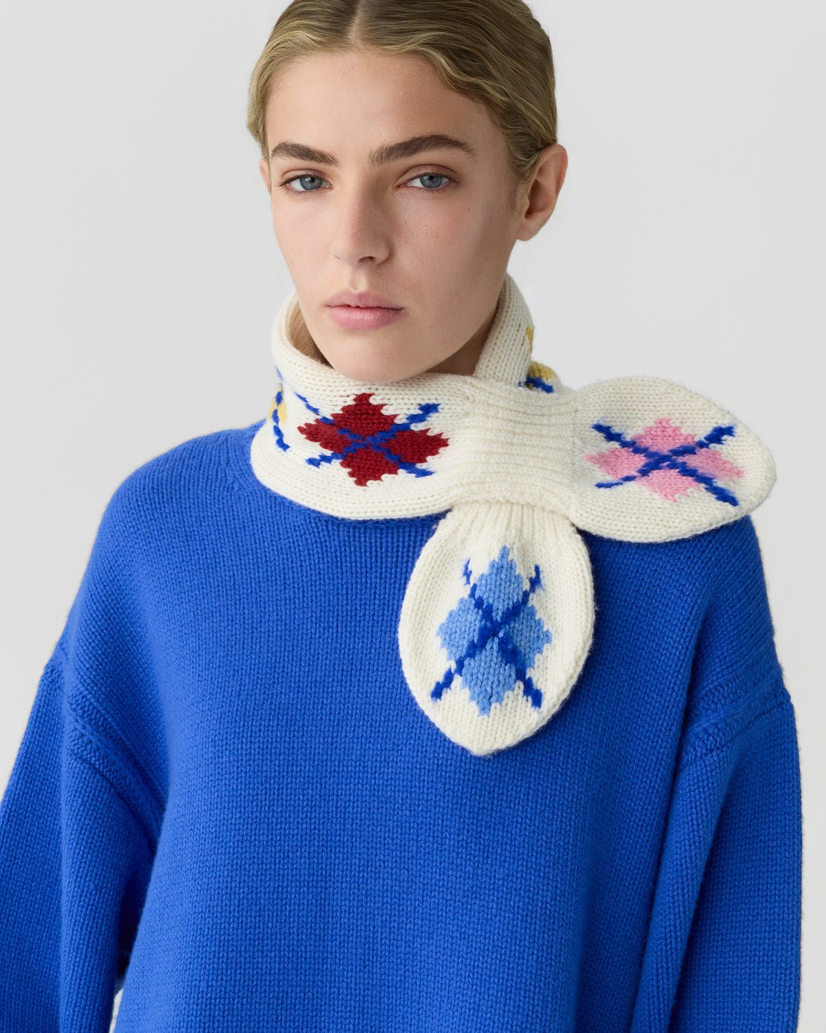 Young woman wearing a bright blue sweater and a white knitted scarf with colorful argyle patterns in red, blue, and pink wrapped around her neck.