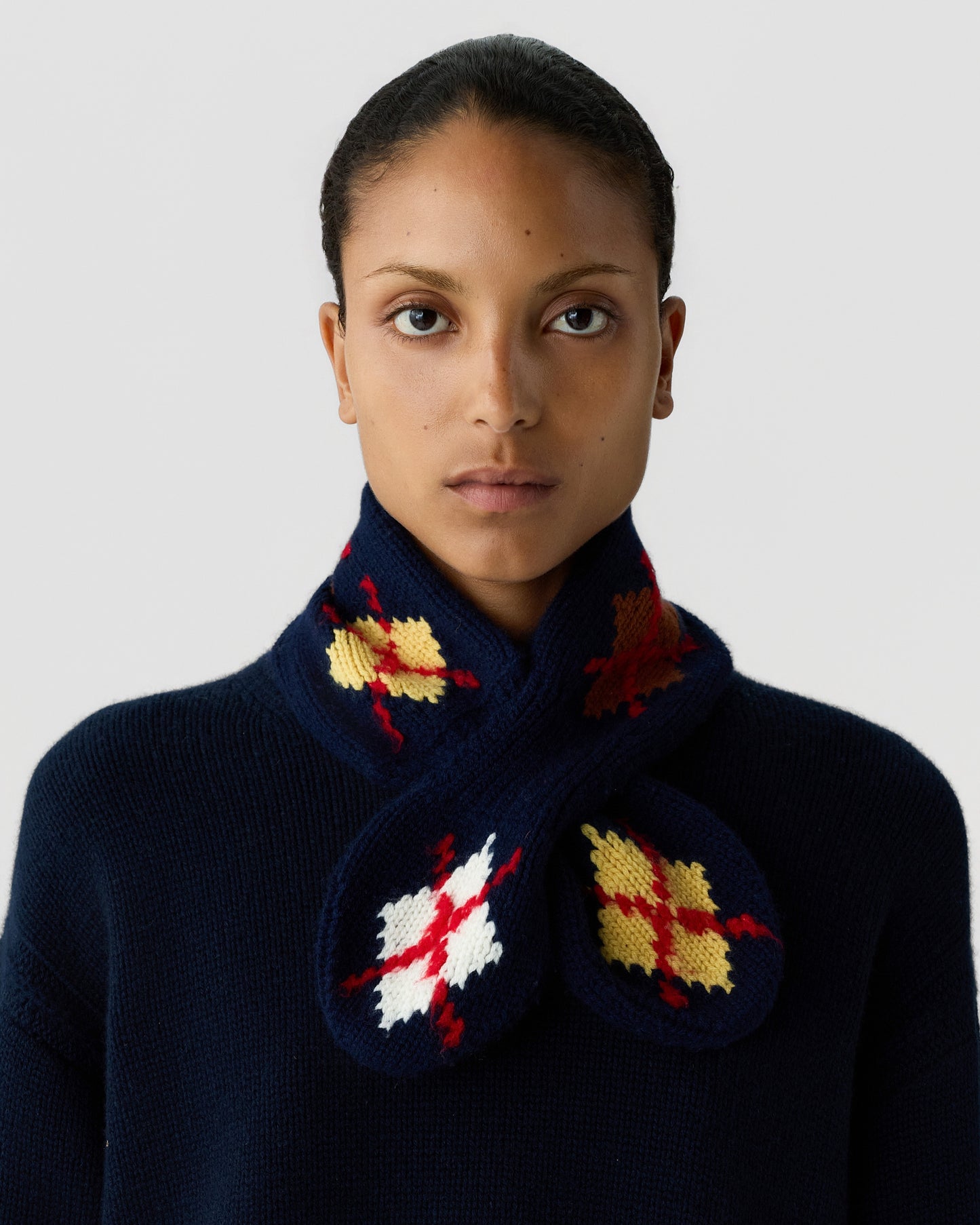 Portrait of a woman wearing a navy blue knitted scarf with yellow, white, and red tartan patterns, styled over a matching navy sweater against a plain background.