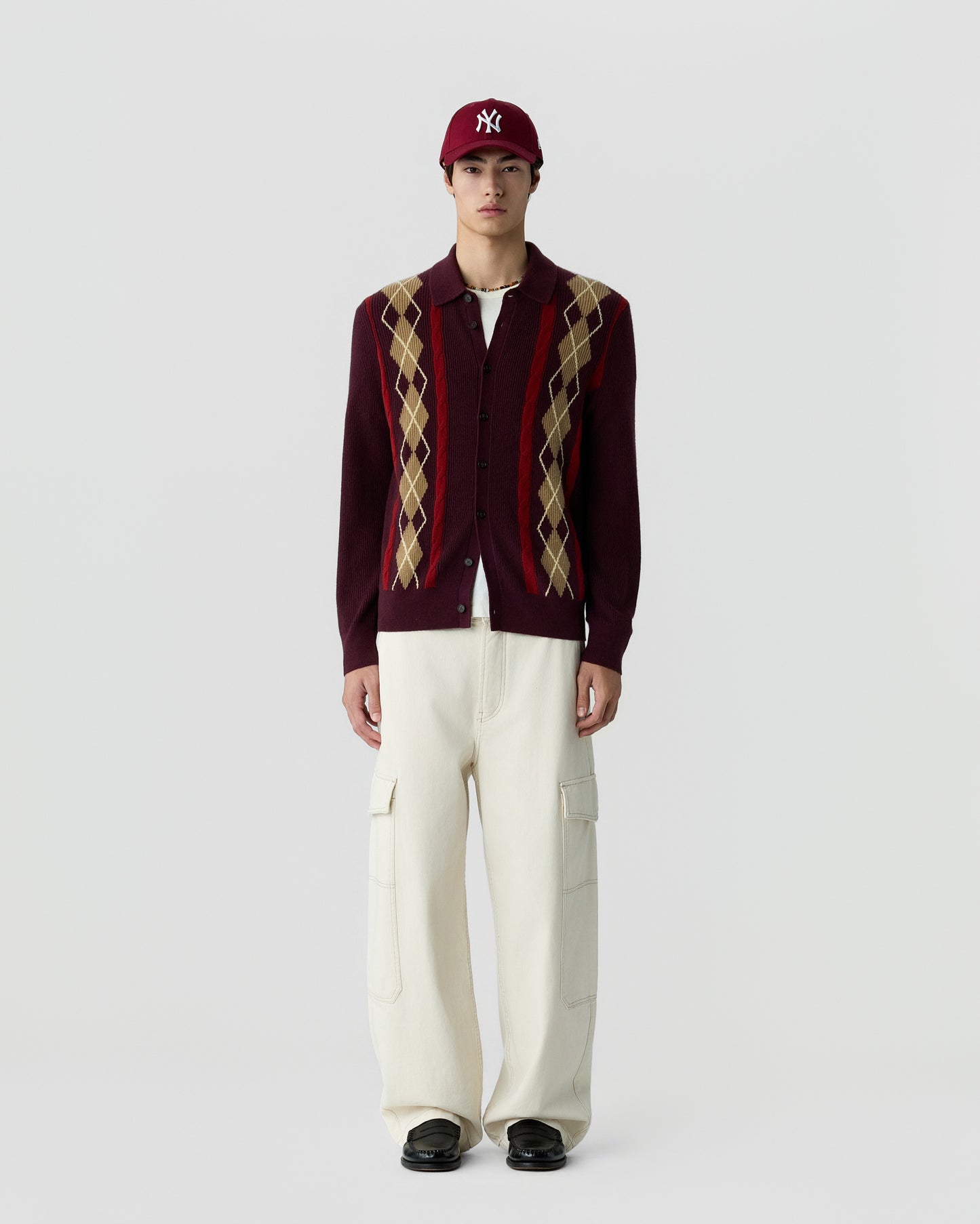 A young man stands against a plain white background wearing a burgundy cardigan with beige and tan argyle patterns, white cargo pants, black shoes, and a dark red baseball cap.