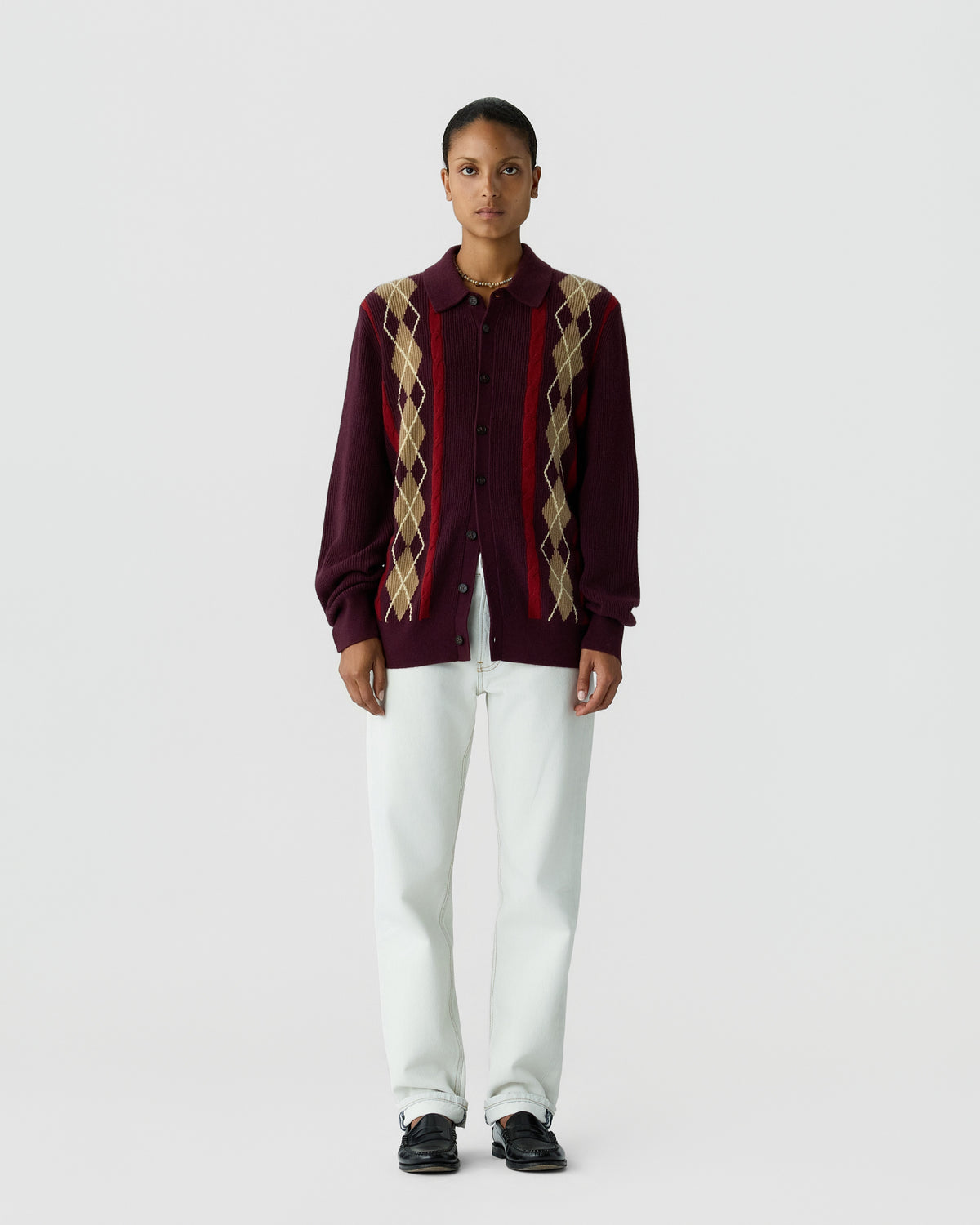 A person stands facing forward wearing a maroon button-up cardigan with beige and red argyle patterns on the front, paired with white pants and black shoes against a plain light gray background.