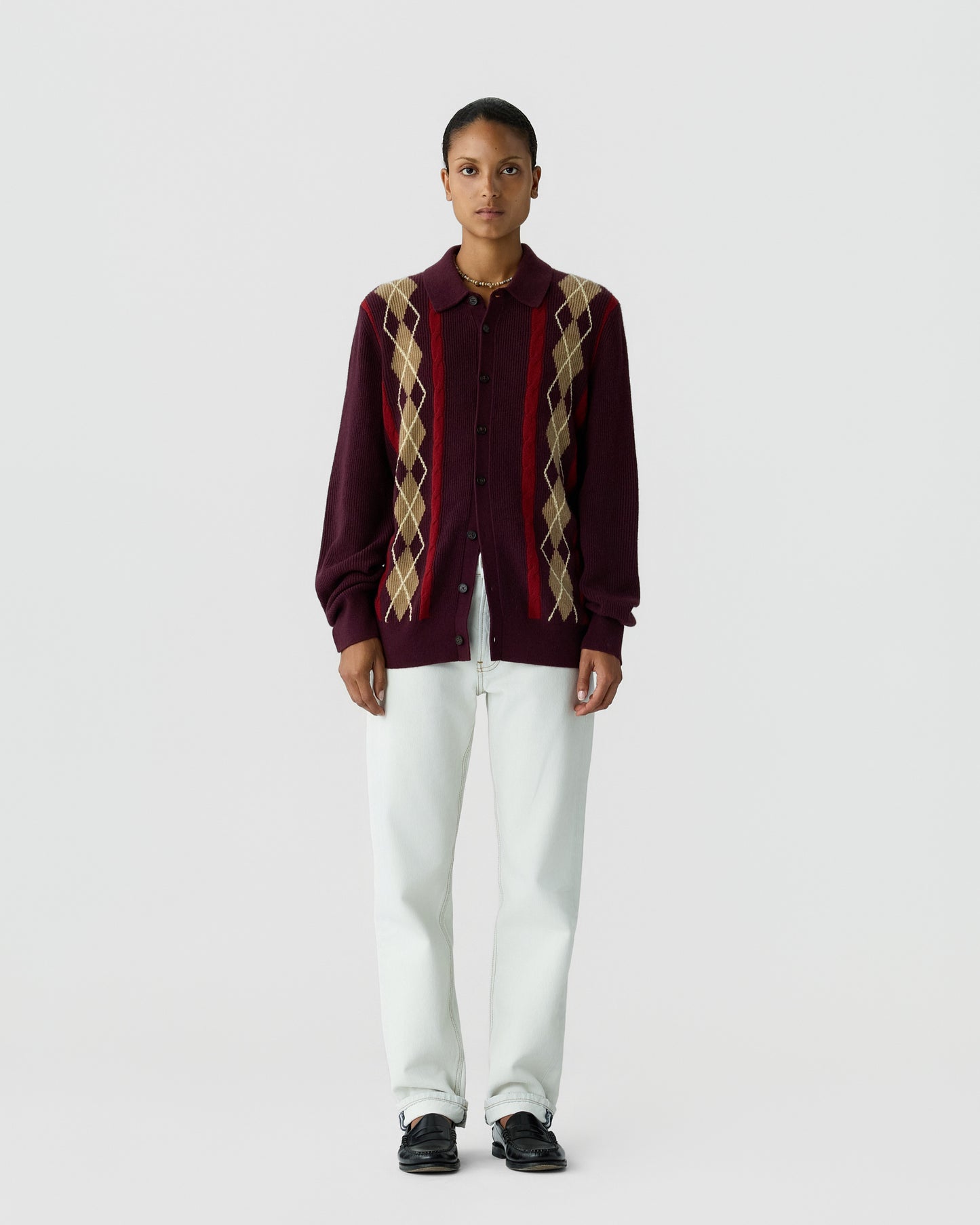 A person stands facing forward wearing a maroon button-up cardigan with beige and red argyle patterns on the front, paired with white pants and black shoes against a plain light gray background.