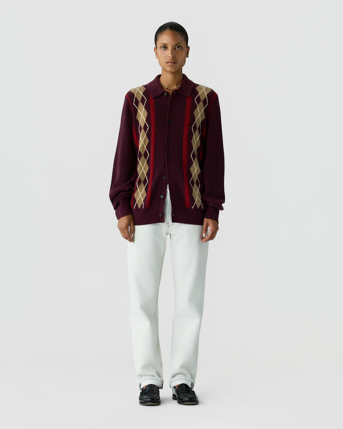 Model standing against a light gray background wearing a burgundy cardigan with argyle stripes over a dark shirt, white trousers and black shoes