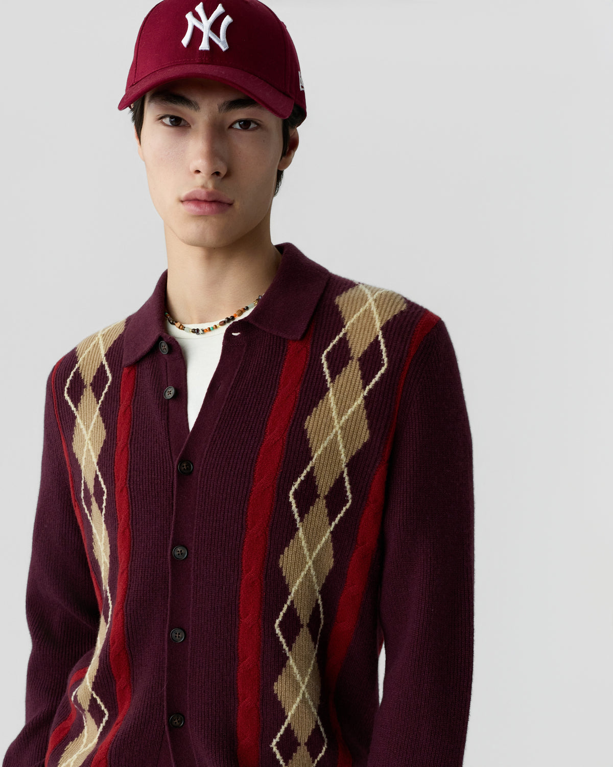Young man wearing a burgundy cardigan with beige and red argyle patterns, a white t-shirt underneath, a beaded necklace, and a maroon baseball cap with a white logo.