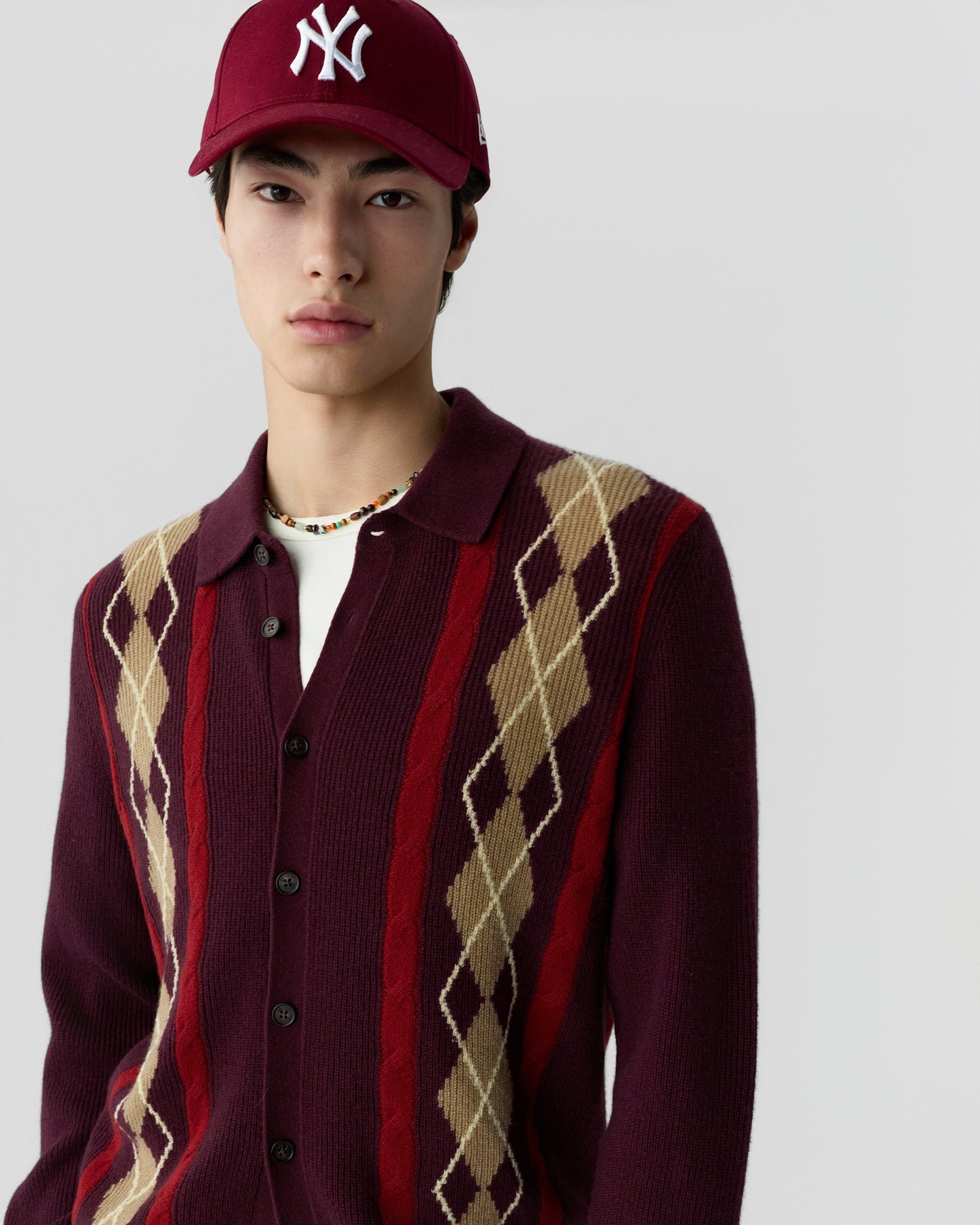 Young man wearing a burgundy cardigan with beige and red argyle patterns, a white t-shirt underneath, a beaded necklace, and a maroon baseball cap with a white logo.