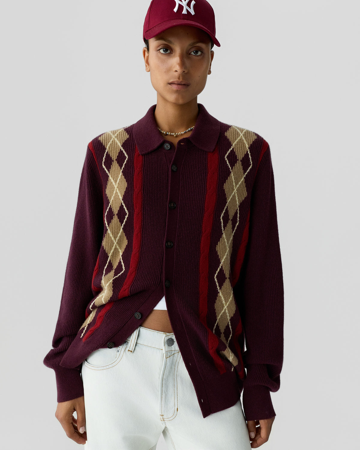 Person wearing a burgundy button-up cardigan with beige and red argyle pattern details, paired with white pants and a burgundy baseball cap with a white logo.