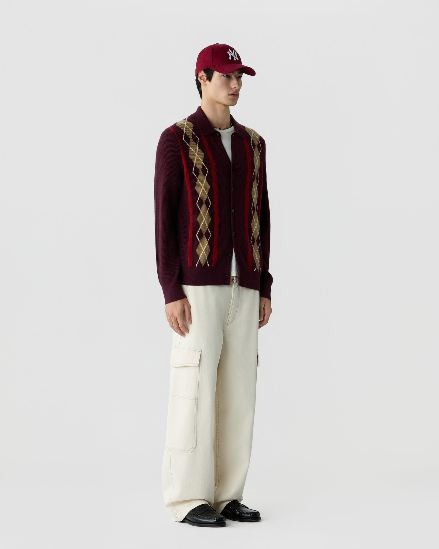 A young man wearing a burgundy argyle-patterned cardigan over a white shirt, cream-colored wide-leg cargo pants, black shoes, and a burgundy baseball cap with a white logo.