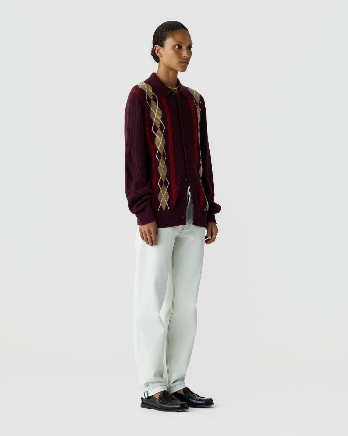 Model wearing a burgundy diamond pattern cardigan with white trousers and black shoes against a light gray studio background