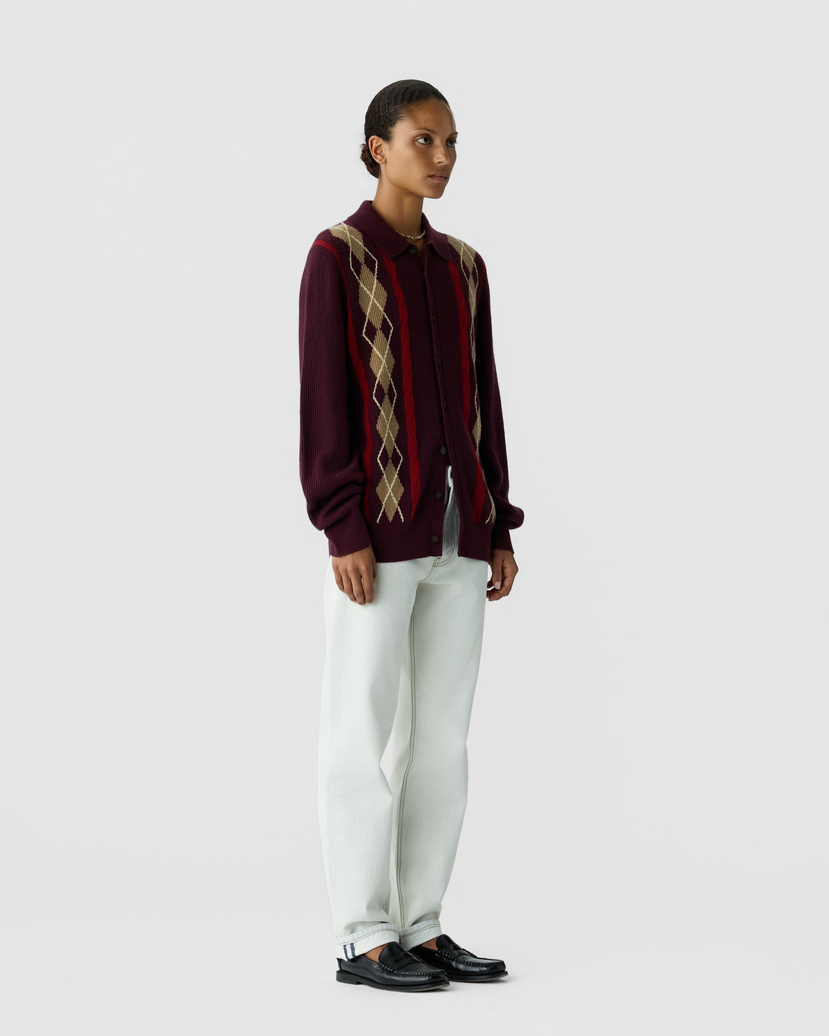 Woman standing wearing a burgundy cardigan with tan and beige argyle pattern, white pants, and black loafers against a plain white background.