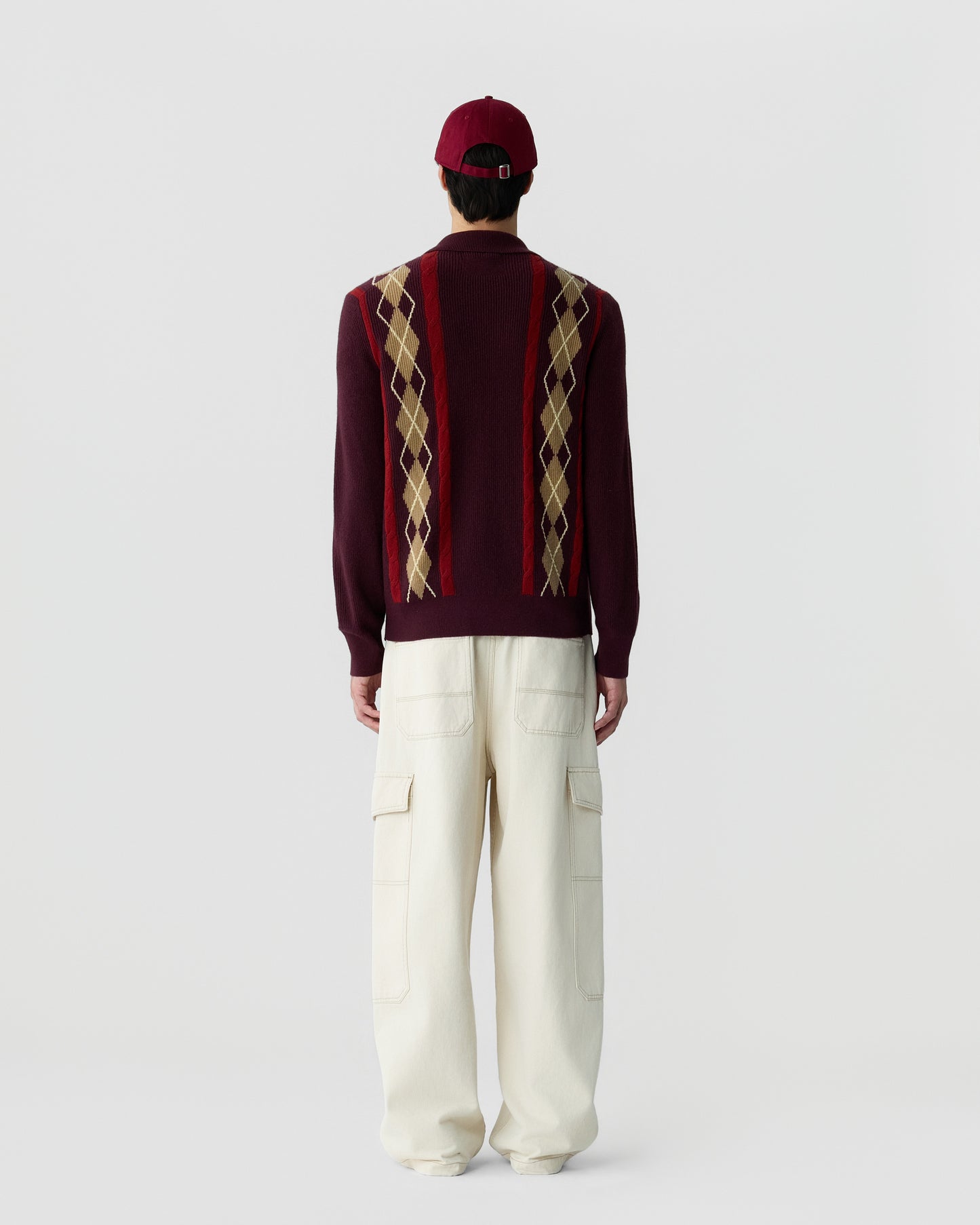 Man wearing a maroon sweater with a beige and red argyle pattern on the back, paired with loose-fitting cream cargo pants and a matching maroon baseball cap, standing against a plain background.