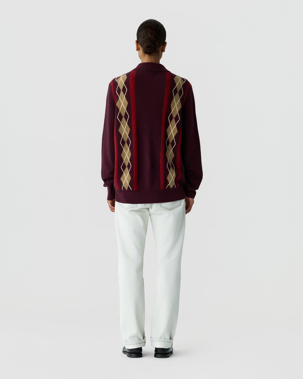 Back view of a person wearing a burgundy sweater with beige and brown argyle stripes, white trousers, and black shoes against a light gray background