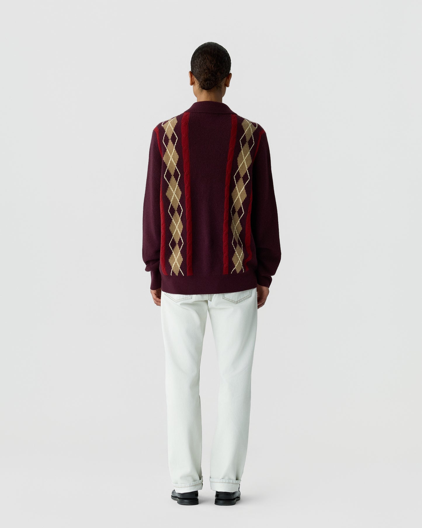 Rear view of a person wearing a burgundy sweater with tan and red argyle patterns down the back, paired with light-colored pants and black shoes against a plain background.