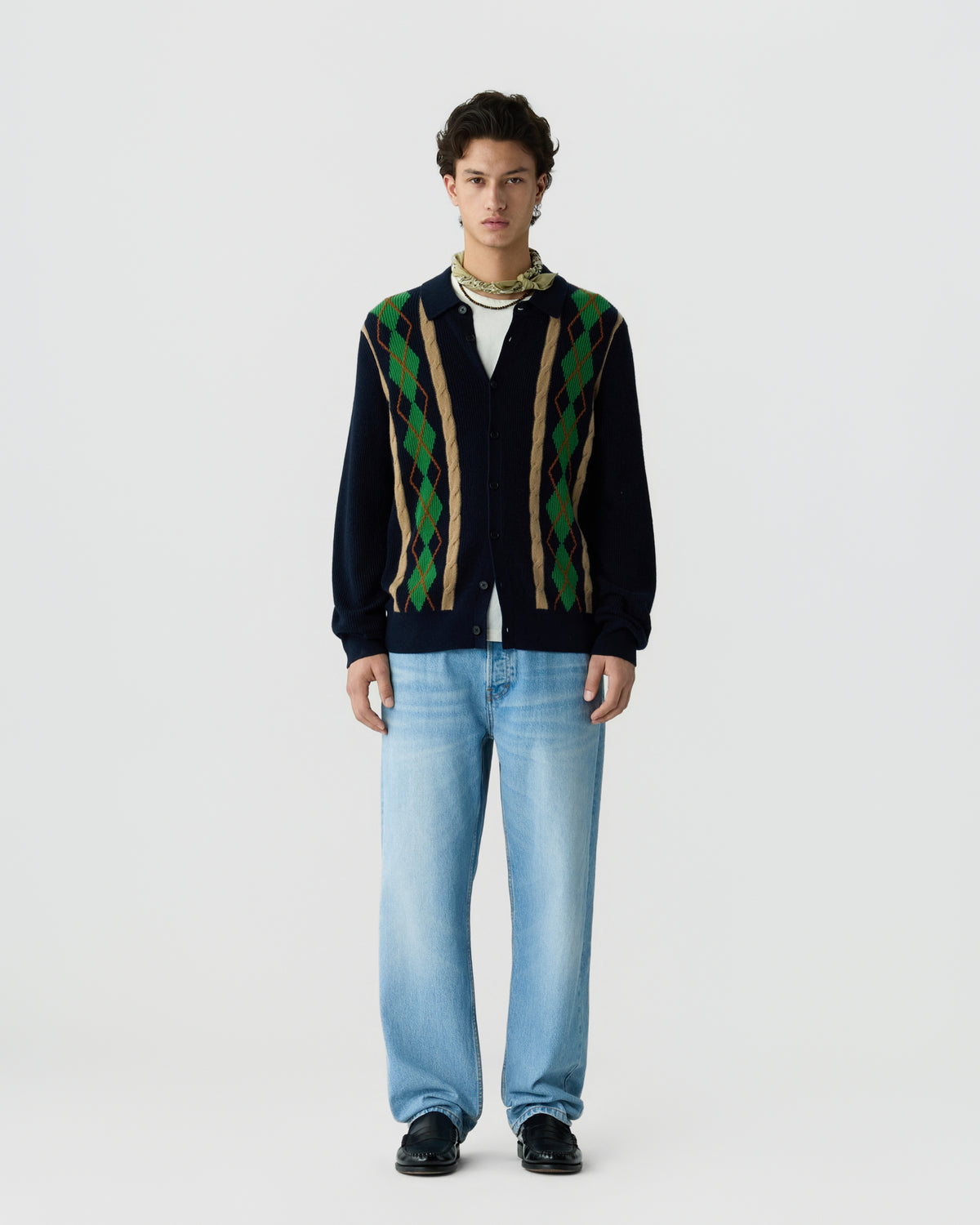Young man wearing a navy cardigan with green and brown argyle patterns over a white shirt, light blue jeans, black shoes, and a patterned scarf around his neck, standing against a plain white background.