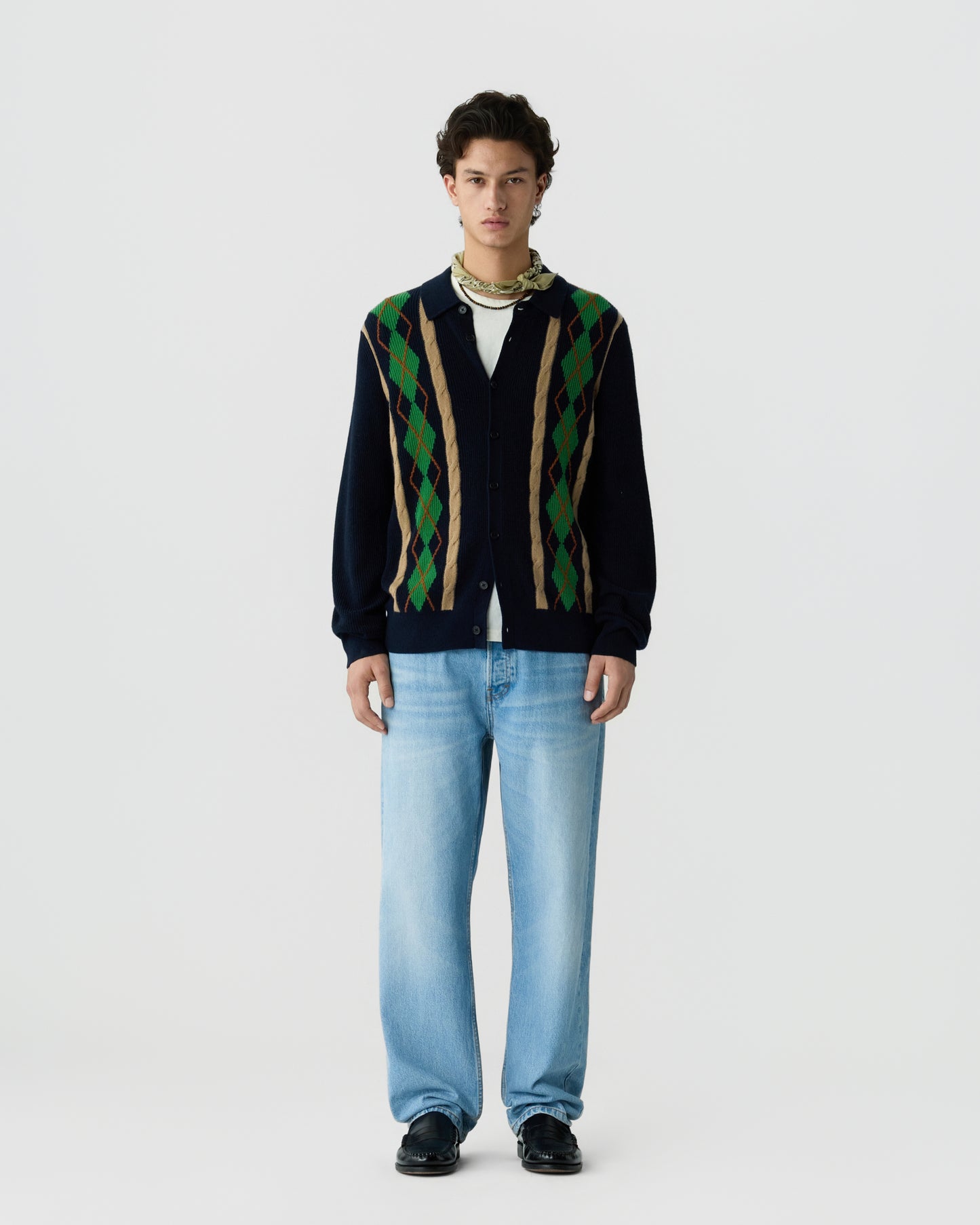 Young man wearing a navy cardigan with green and brown argyle patterns over a white shirt, light blue jeans, black shoes, and a patterned scarf around his neck, standing against a plain white background.
