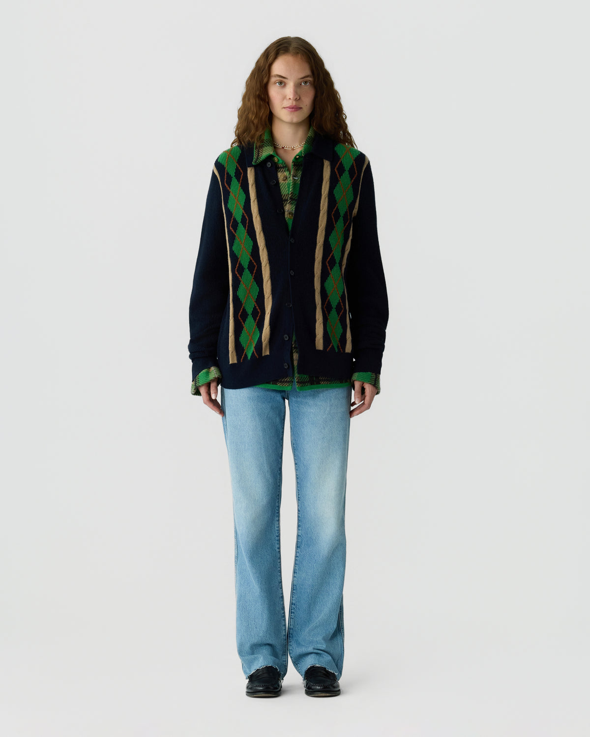 Model wearing a dark navy cardigan with green and brown argyle panels over a green collared shirt with light blue jeans and black shoes