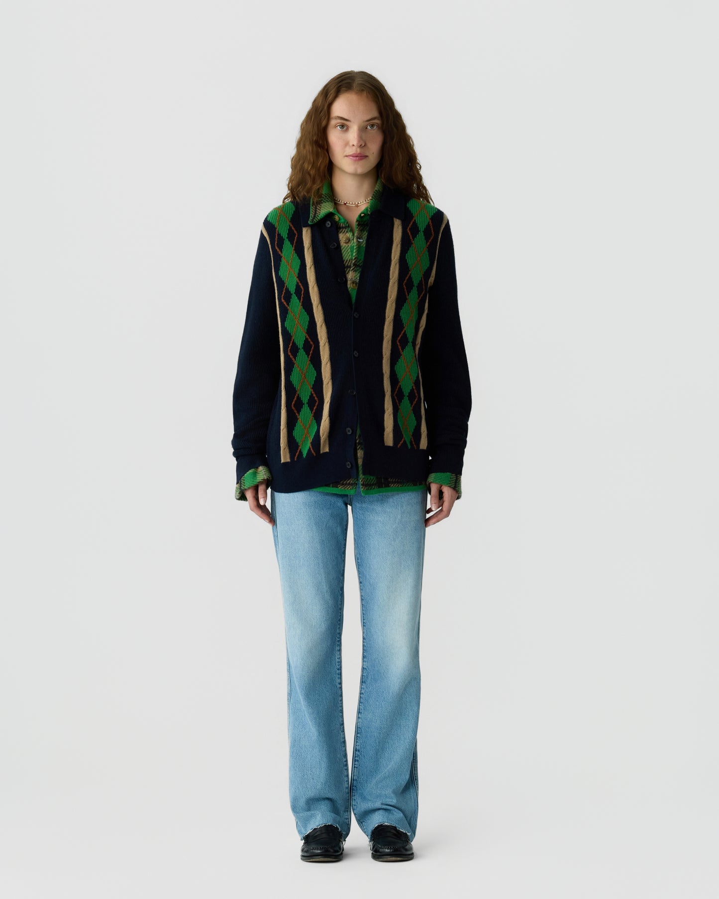Woman with long curly hair wearing a dark cardigan with green and beige argyle pattern over a green plaid shirt, light blue jeans, and black shoes standing against a plain white background.