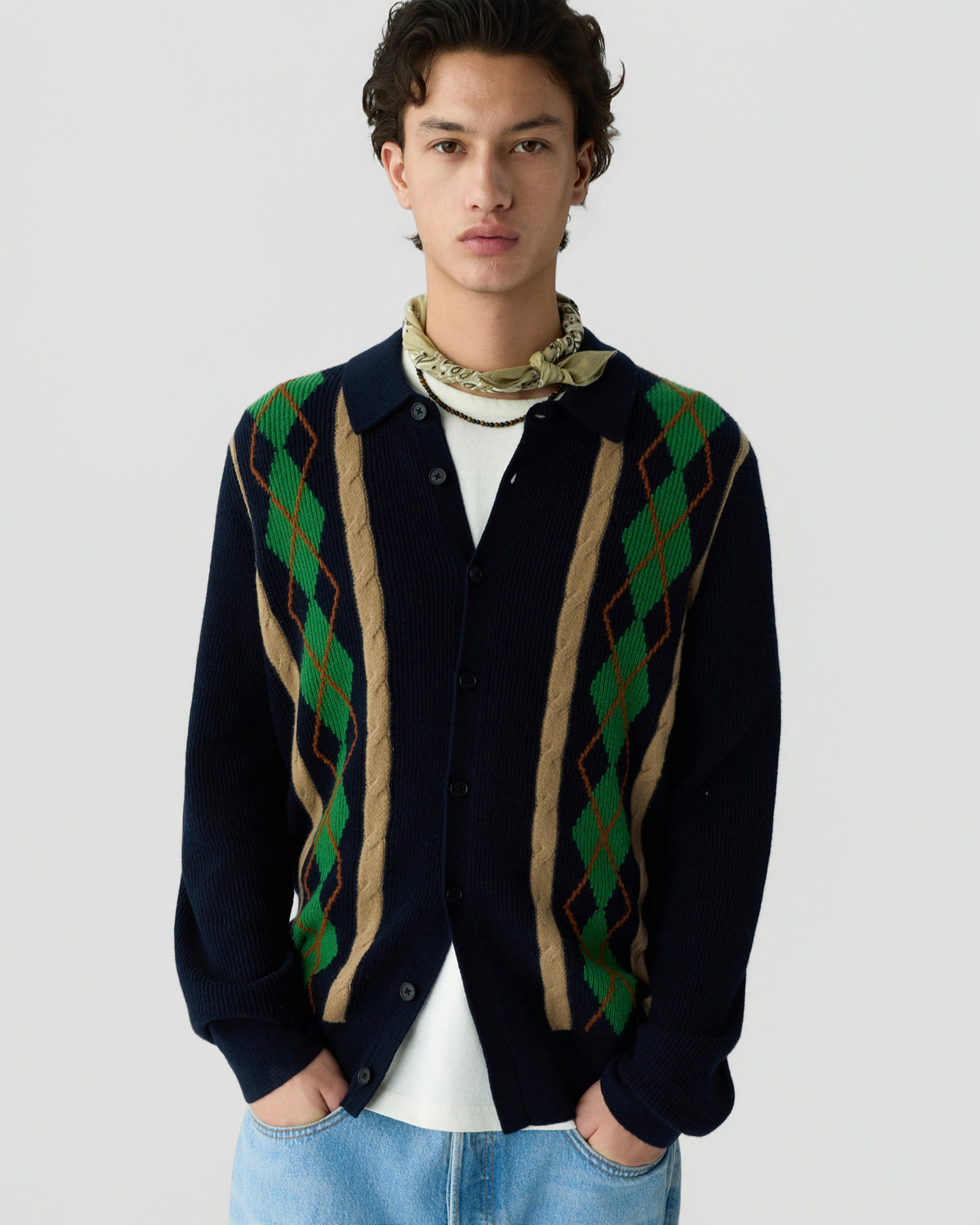 Young man wearing a navy blue cardigan with green and beige argyle patterns over a white shirt, light blue jeans, and a patterned beige neck scarf.