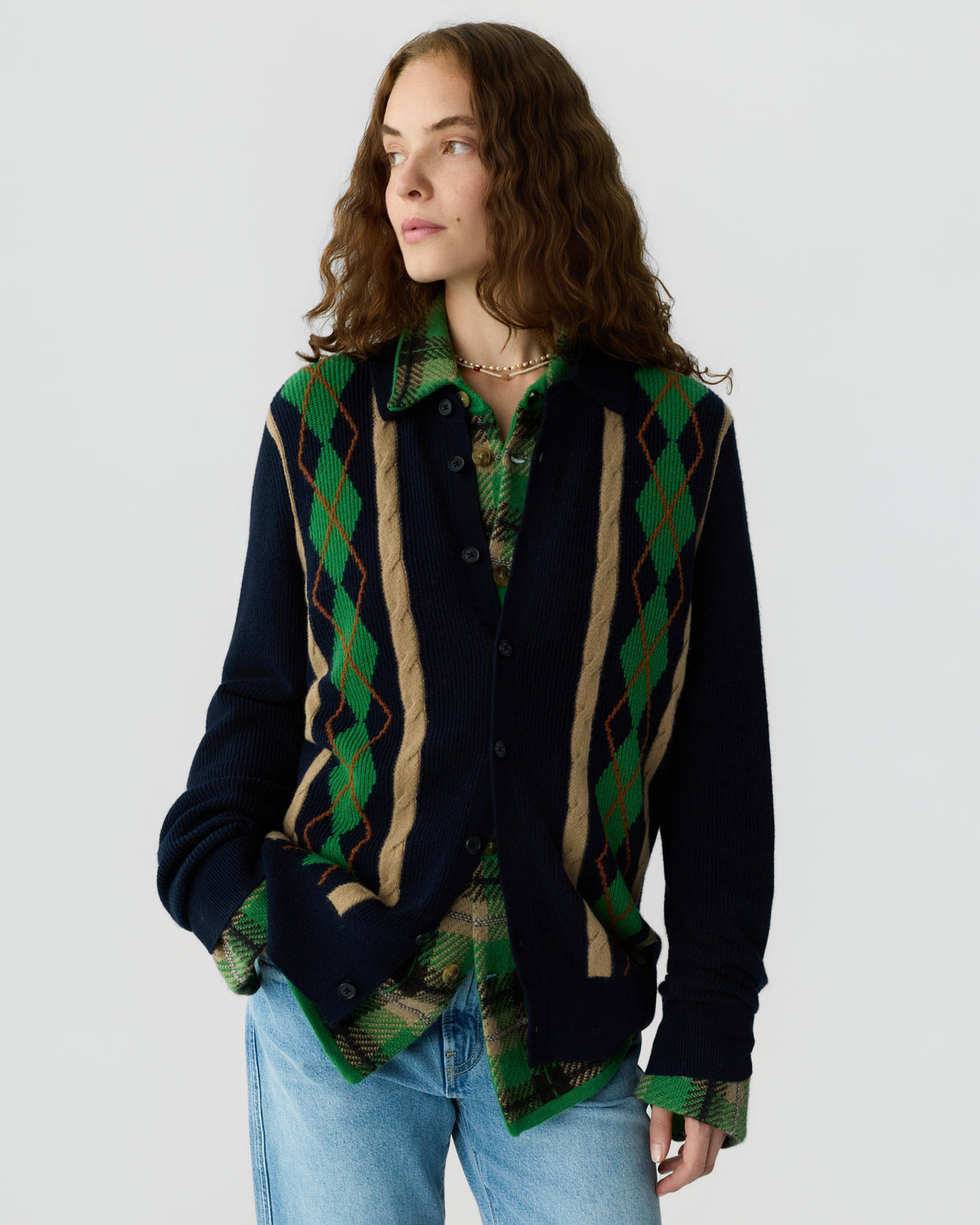 Woman with wavy brown hair wearing a navy blue cardigan with beige and green argyle stripes over a green plaid shirt and light blue jeans, standing against a plain white background.