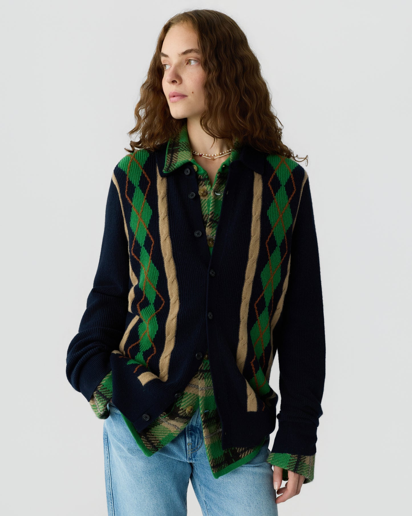 Woman with wavy brown hair wearing a navy blue cardigan with beige and green argyle stripes over a green plaid shirt and light blue jeans, standing against a plain white background.