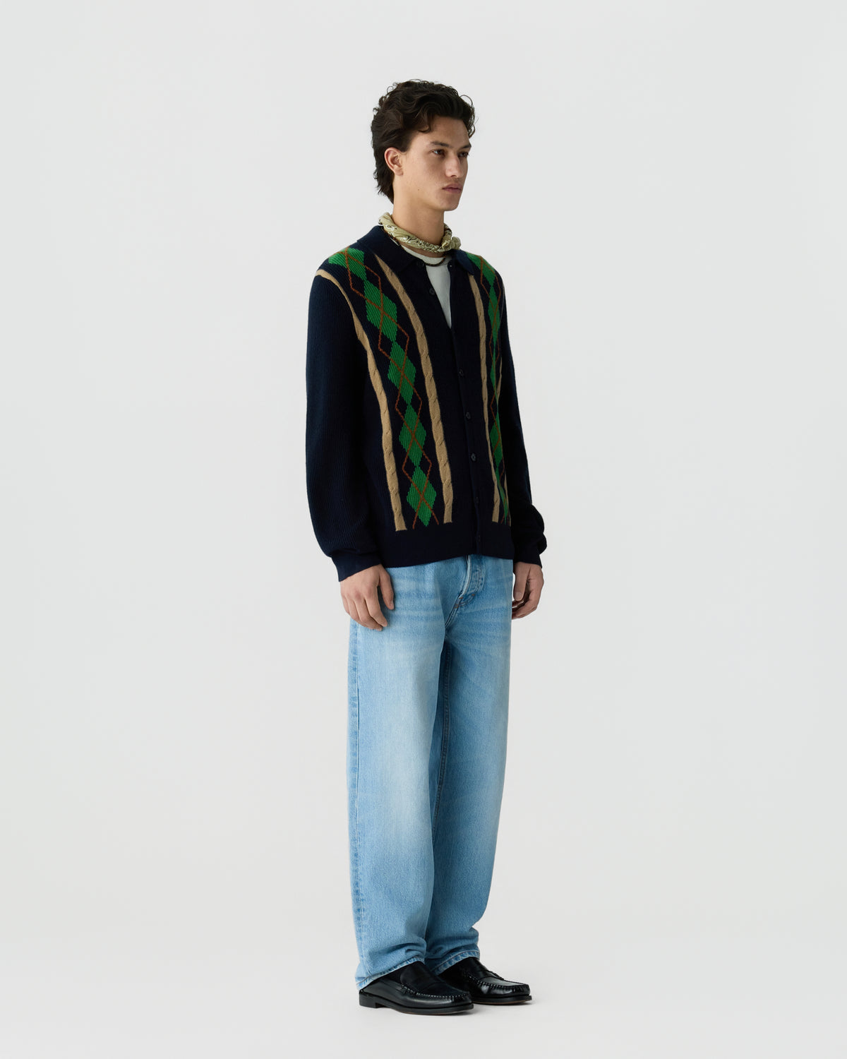Young man wearing a navy blue cardigan with green and brown argyle pattern, light blue jeans, black shoes, and a beige scarf around his neck, standing against a plain white background.