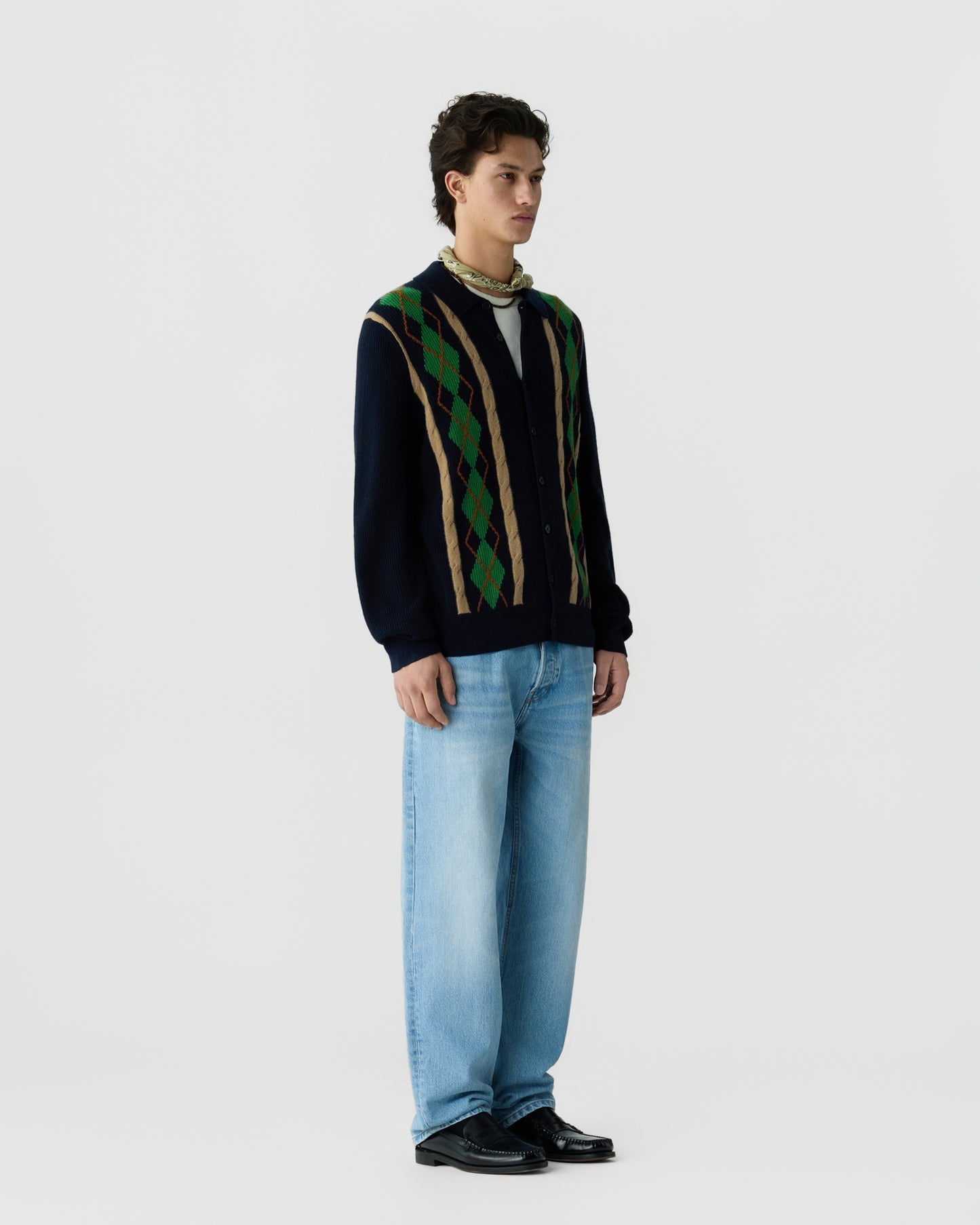 Young man wearing a navy blue cardigan with green and brown argyle pattern, light blue jeans, black shoes, and a beige scarf around his neck, standing against a plain white background.