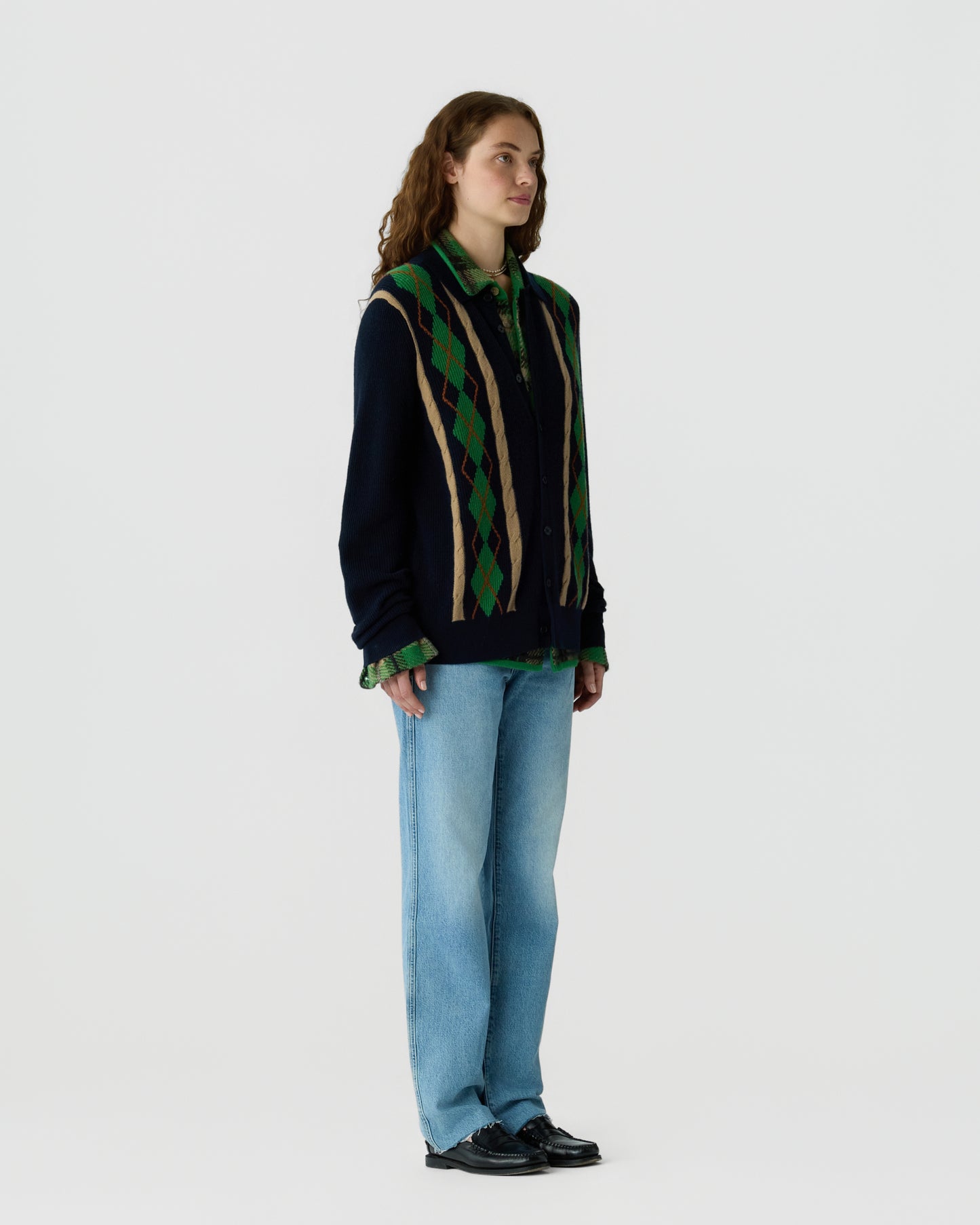 Woman with long curly hair wearing a navy sweater with green and beige argyle pattern over a green plaid shirt, light blue jeans, and black shoes standing against a plain background.