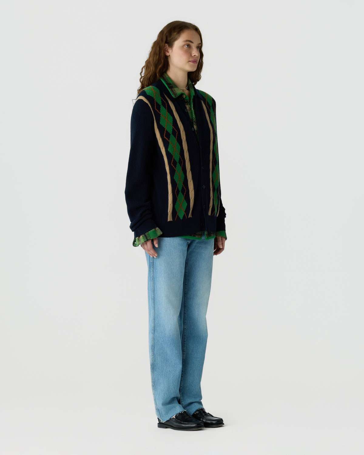 Person wearing a navy patterned knit sweater with green accents, light blue jeans, and black loafers against a light gray background